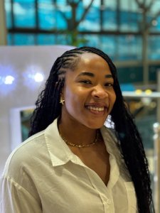 A young woman with shoulder-length brown hair, wearing a beige sleeveless top and layered necklaces, standing outdoors in a park with trees and buildings in the background.