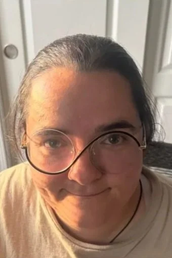Close-up of a person with long gray hair, glasses, and a slight smile, indoors.