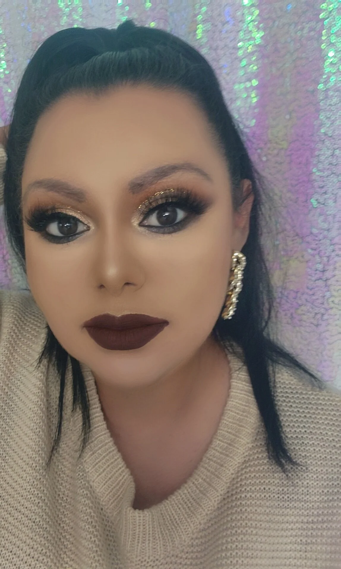 A woman with dark hair styled back, wearing bold makeup with gold and black eyeshadow, dark lipstick, gold earrings, and a beige knit sweater, lying on a sparkly, iridescent background.