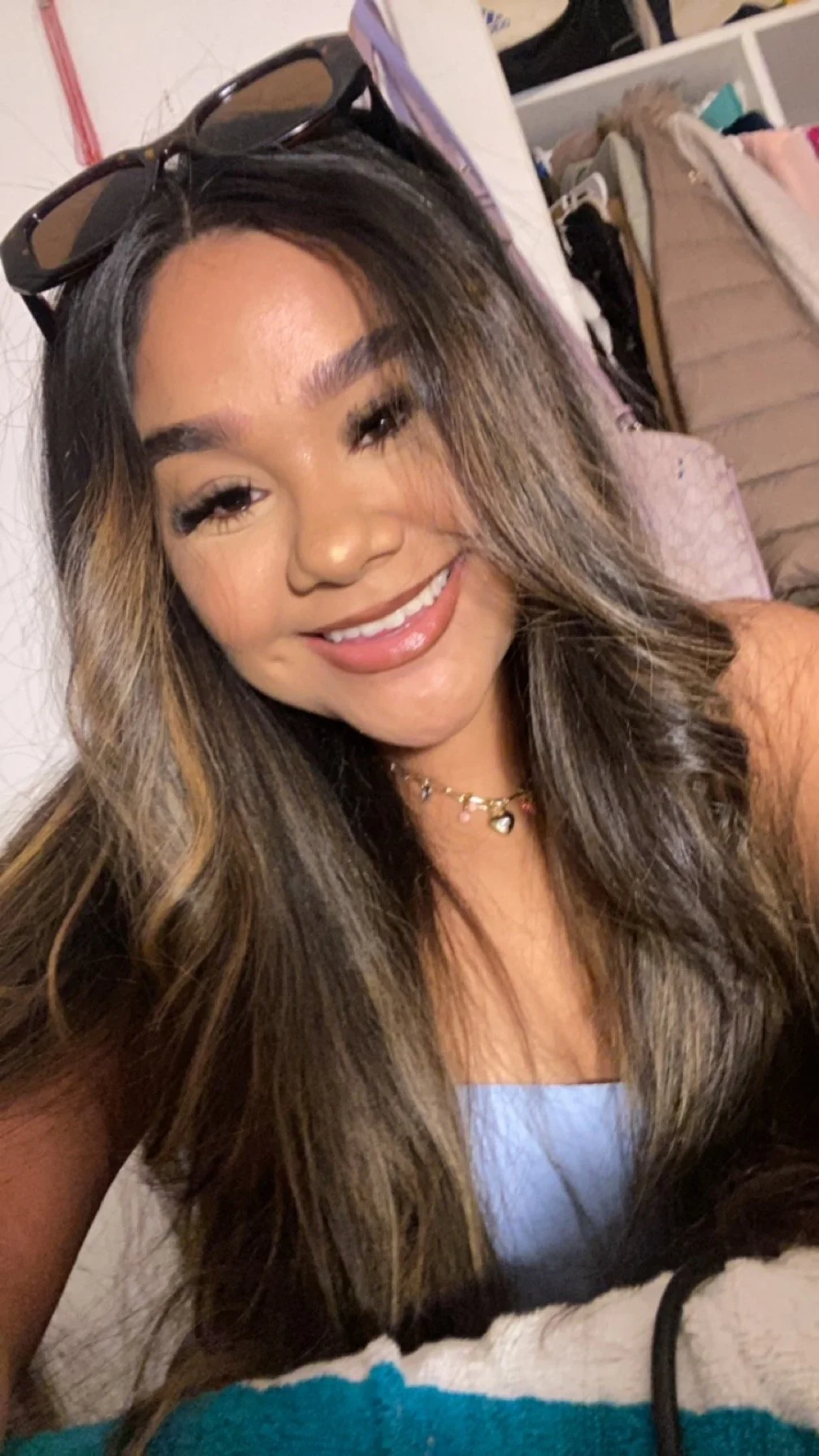 A woman smiling, wearing sunglasses on her head and a necklace, with long wavy hair, indoors with shelves and clothing in the background.