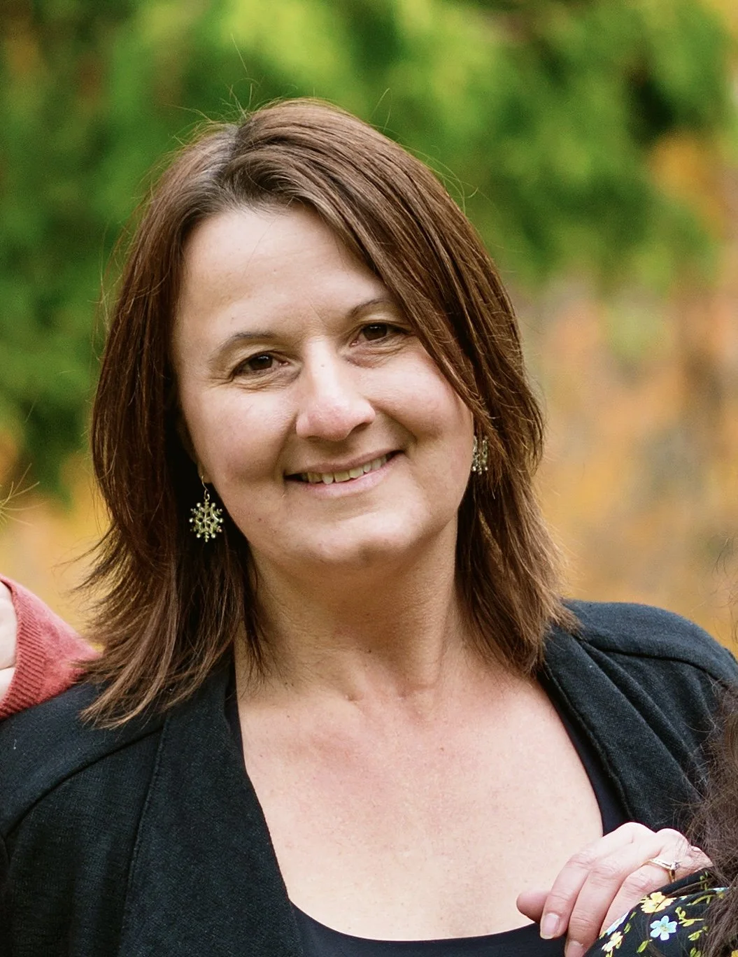 A woman with shoulder-length brown hair smiling outdoors, wearing snowflake-shaped earrings and a black top with a dark blazer, with a blurred background of greenery.