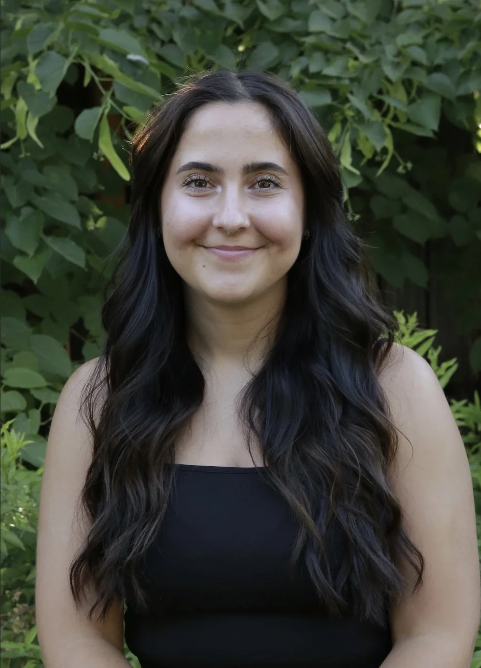 A young woman with long black wavy hair smiling outdoors in front of green foliage, wearing a black sleeveless top.