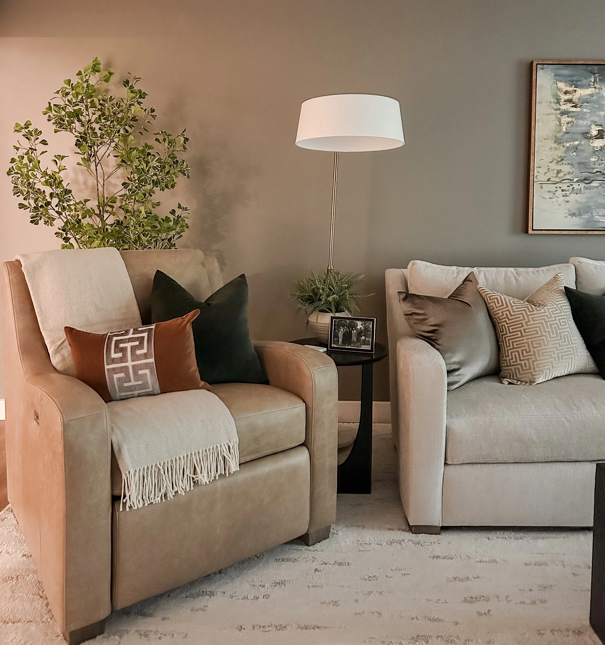 Living room with two beige sofas, decorative pillows, a small black side table with a framed photo, a green plant, a tall floor lamp, and a framed painting on the wall.
