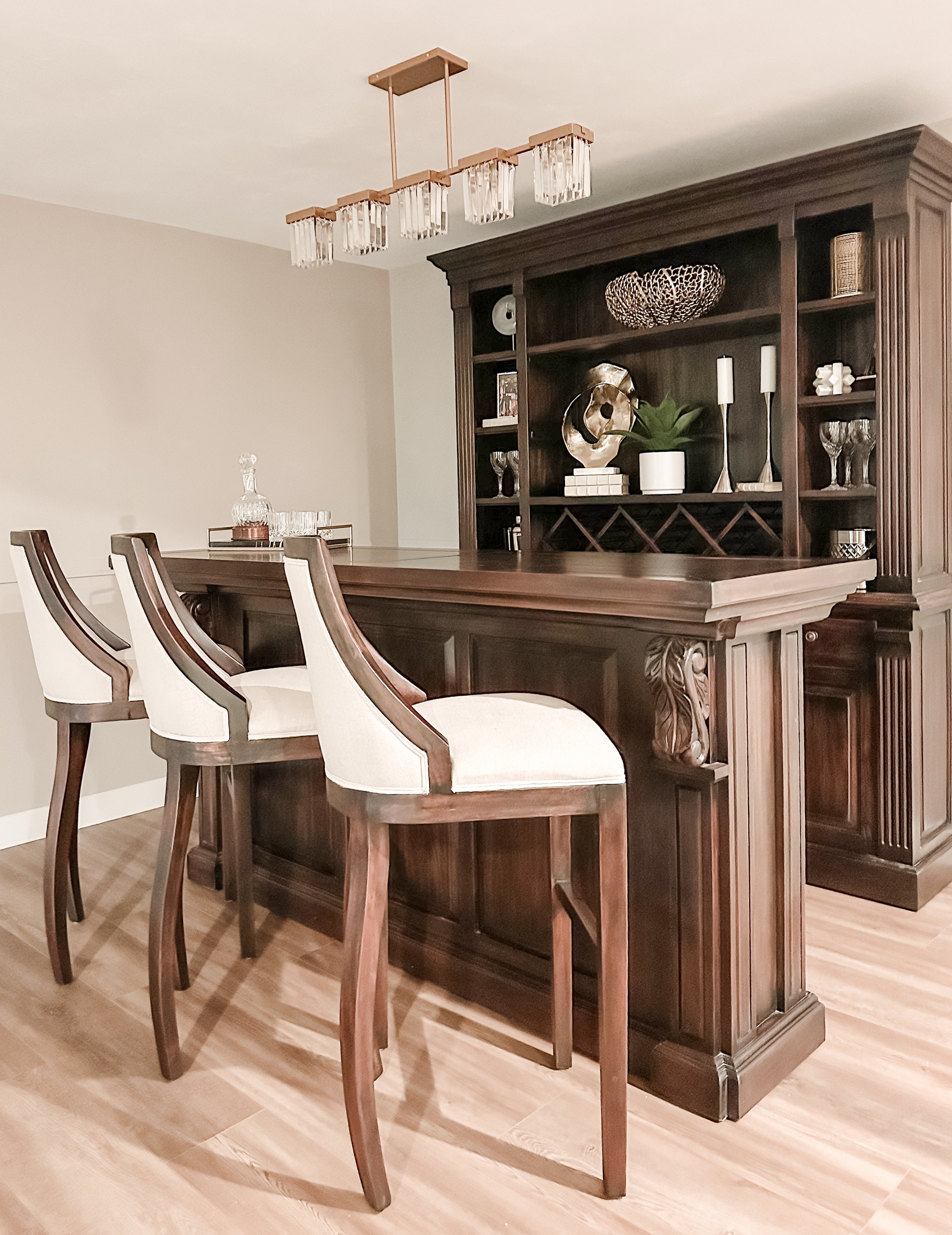 A home bar area with a dark wood counter and matching shelving unit, three white cushioned bar stools, and decorative items like a glass decanter, candles, and sculptures.