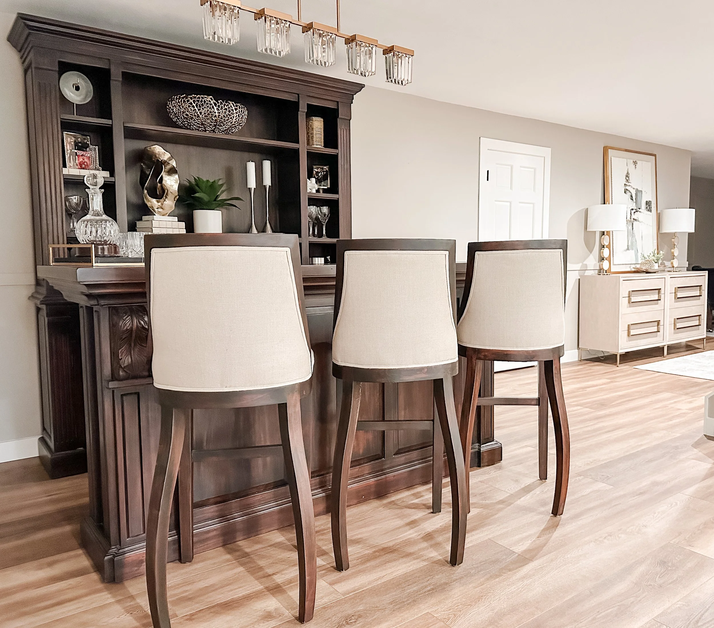 A dining area with three upholstered bar stools in front of a dark wooden bar with decorative items, a wood cabinet with lamps and artwork, and a hardwood floor.