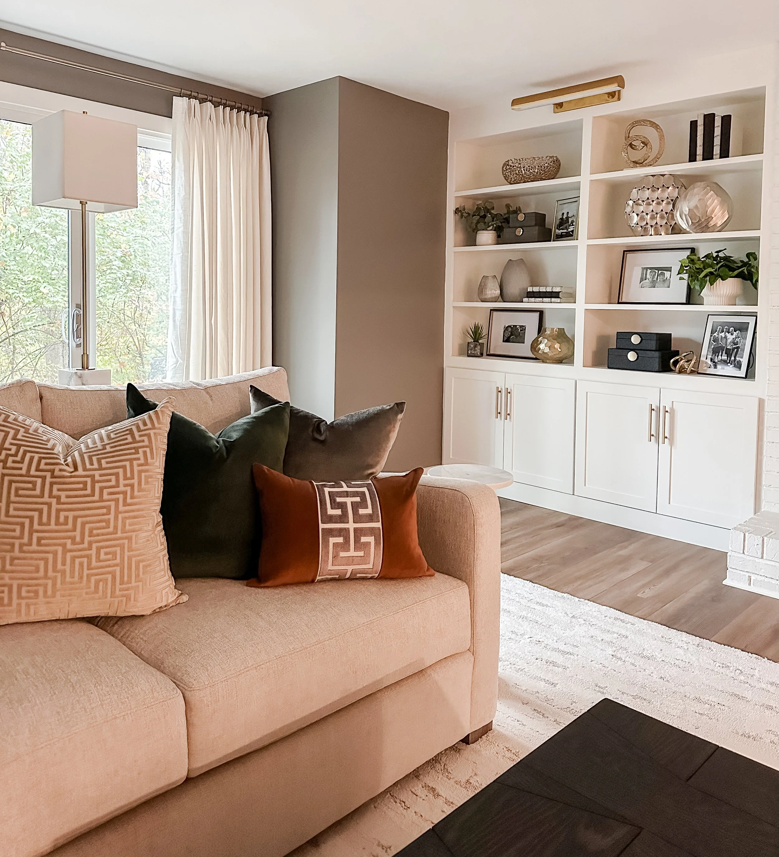 A cozy living room with a beige sofa adorned with decorative pillows, near a window with white curtains. A white bookshelf with decorative items and framed photos is against the wall, with a dark wood coffee table in the foreground and a neutral-colo