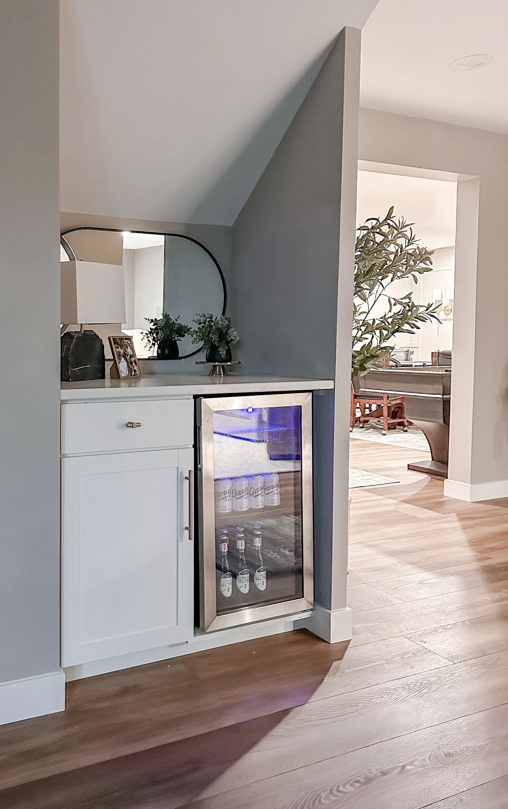 A built-in drink station with a small refrigerator containing bottled drinks, located against a wall with decorative items and a mirror on top, in a modern home interior.