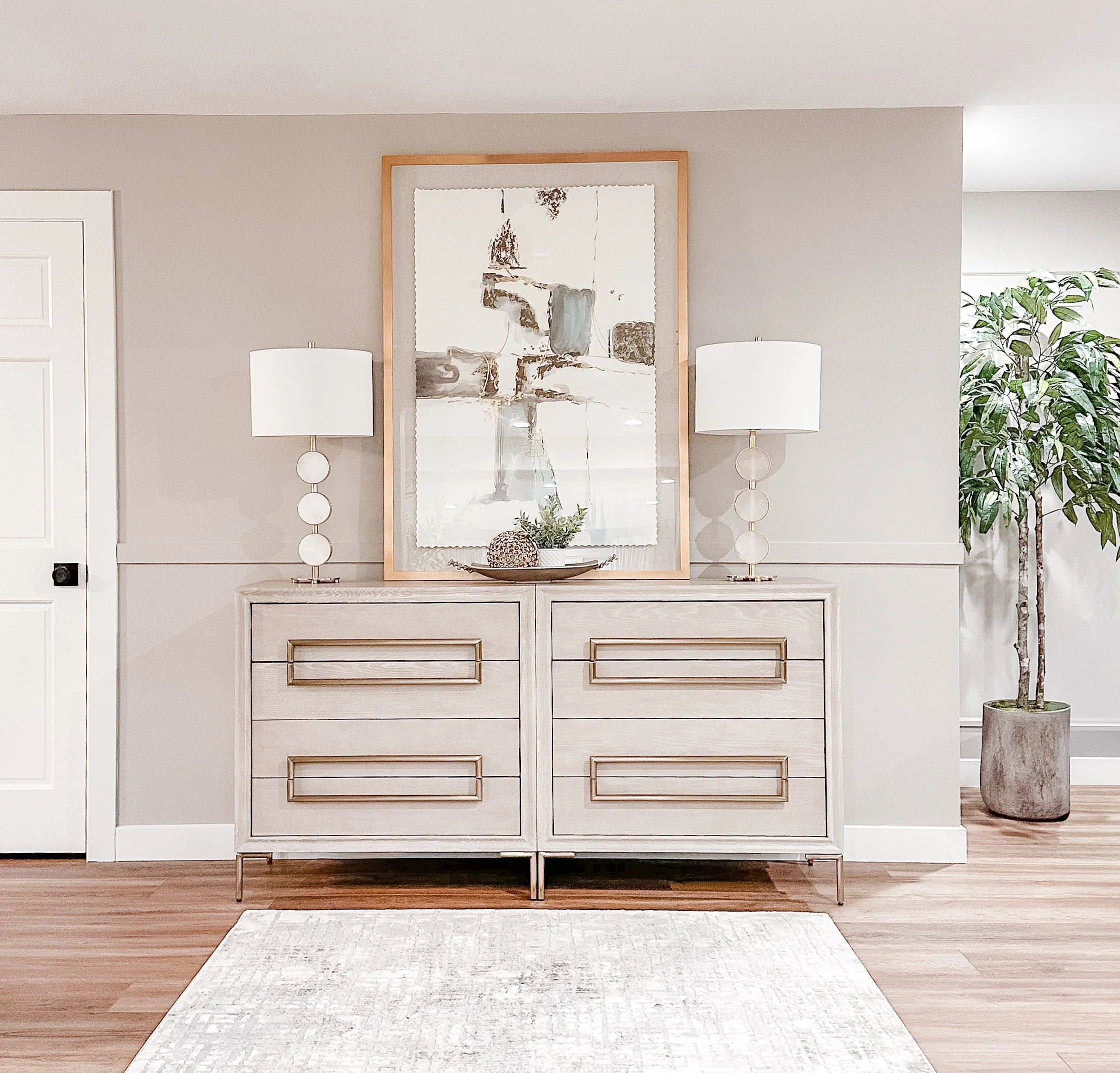 Living room entryway with a light-colored dresser topped with two modern lamps, a framed abstract art piece, and decorative objects, with a large potted plant to the side.