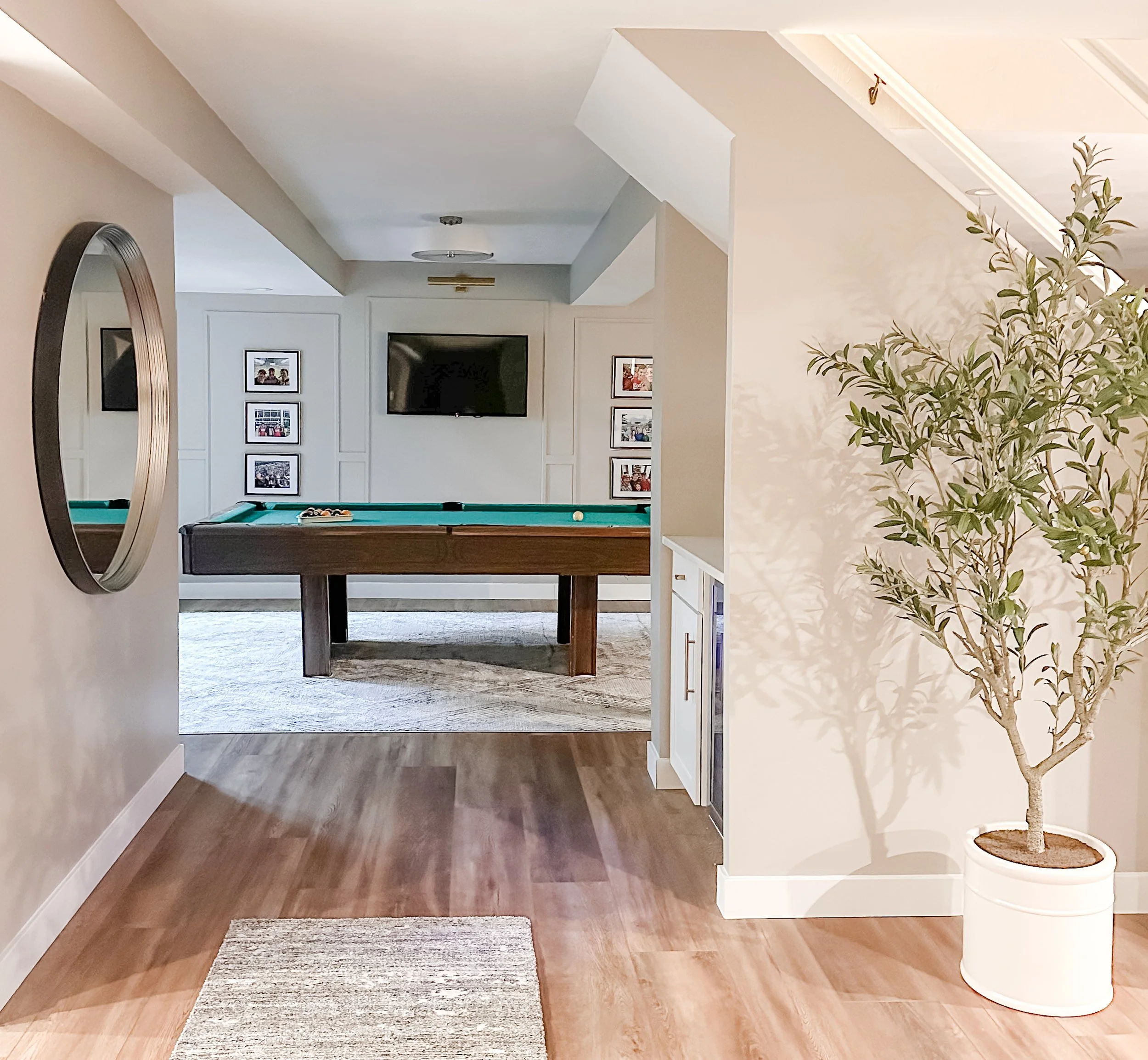 A room with a pool table in the center, a wall-mounted TV above it, framed pictures on the wall, a round mirror on the left wall, and a tall potted plant on the right.