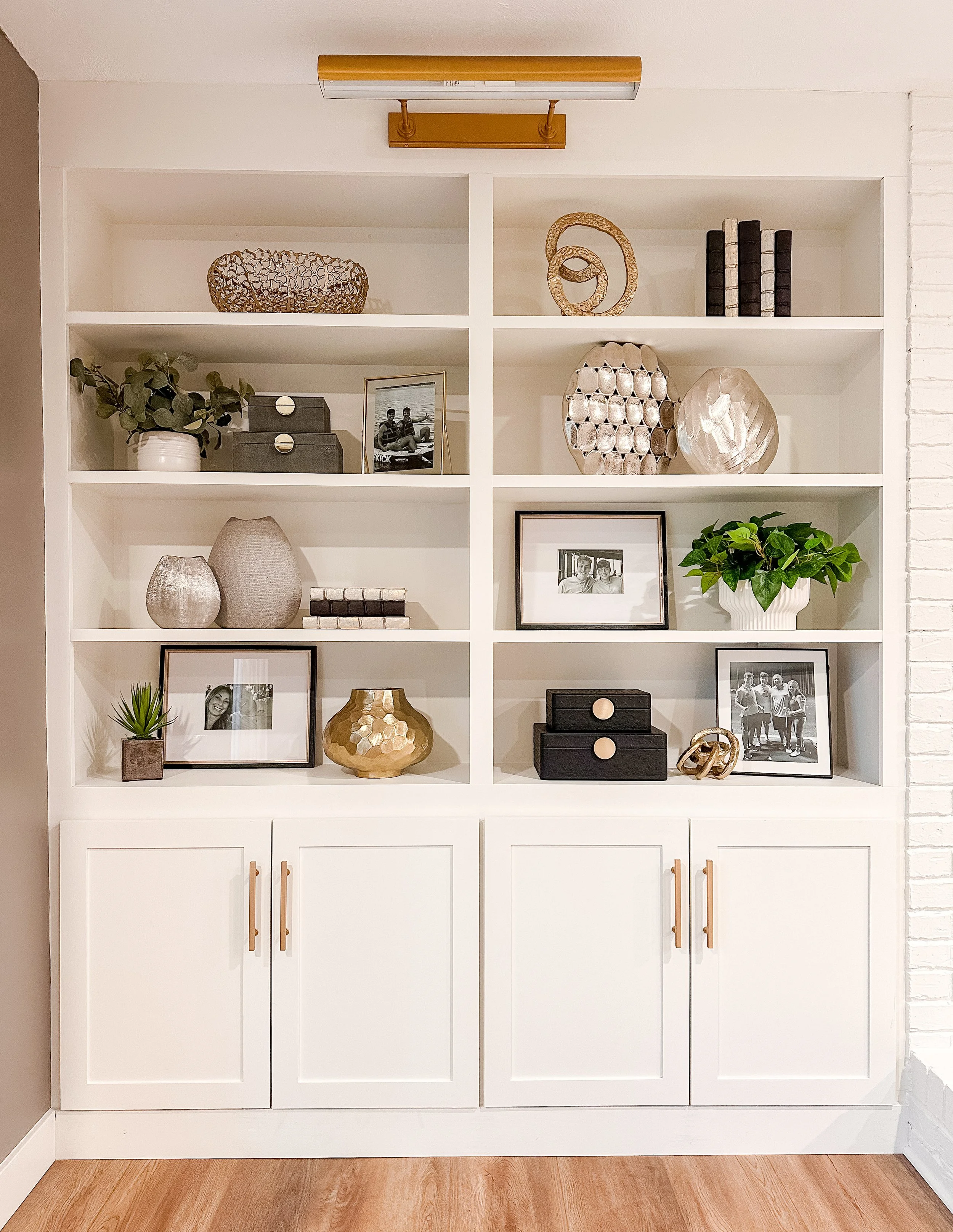 White built-in bookshelf with decorative items, framed photos, and potted plants, beneath a ceiling light fixture.