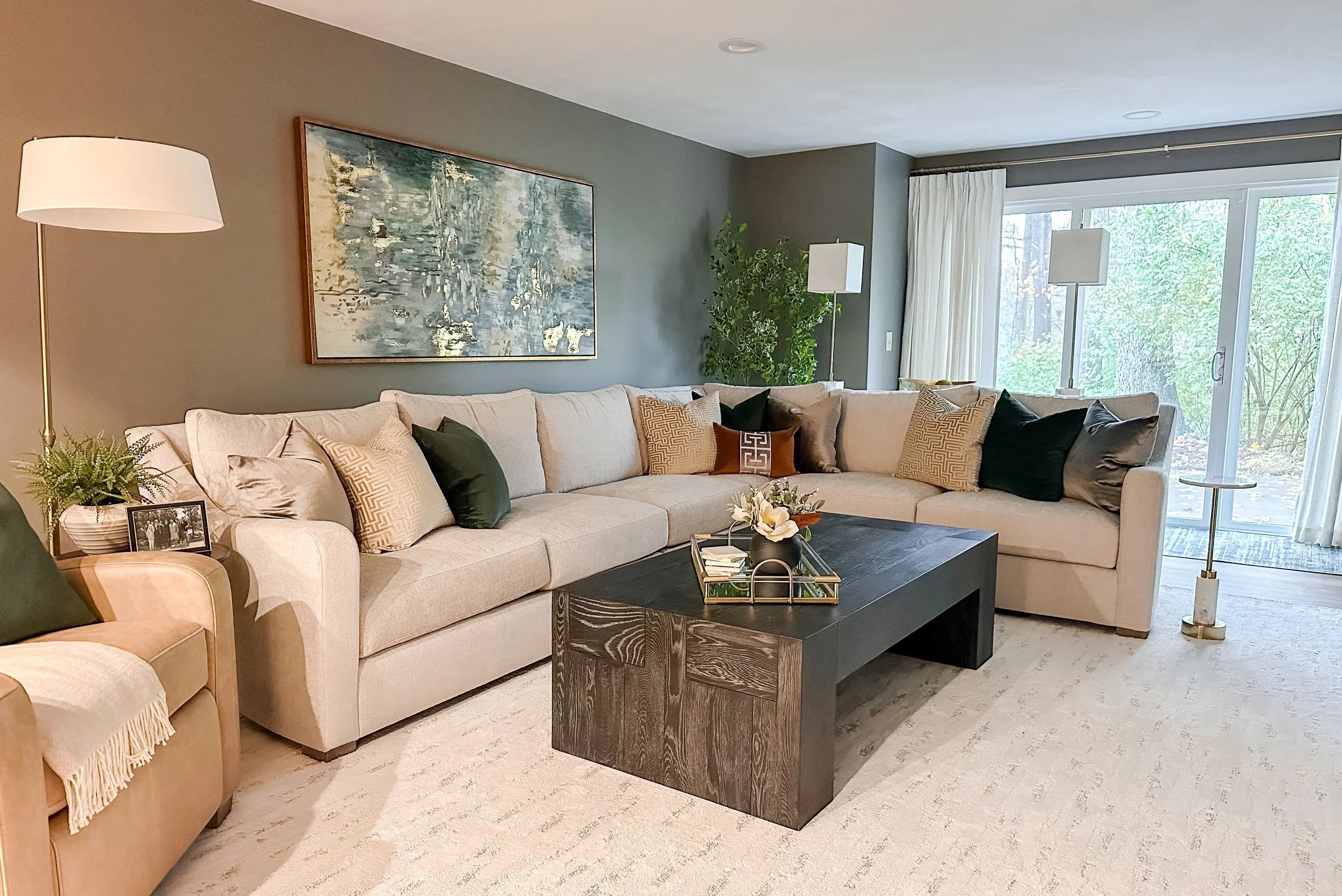 Living room with beige sectional sofa, black coffee table, abstract painting, floor lamps, potted plant, and sliding glass door with white curtains.