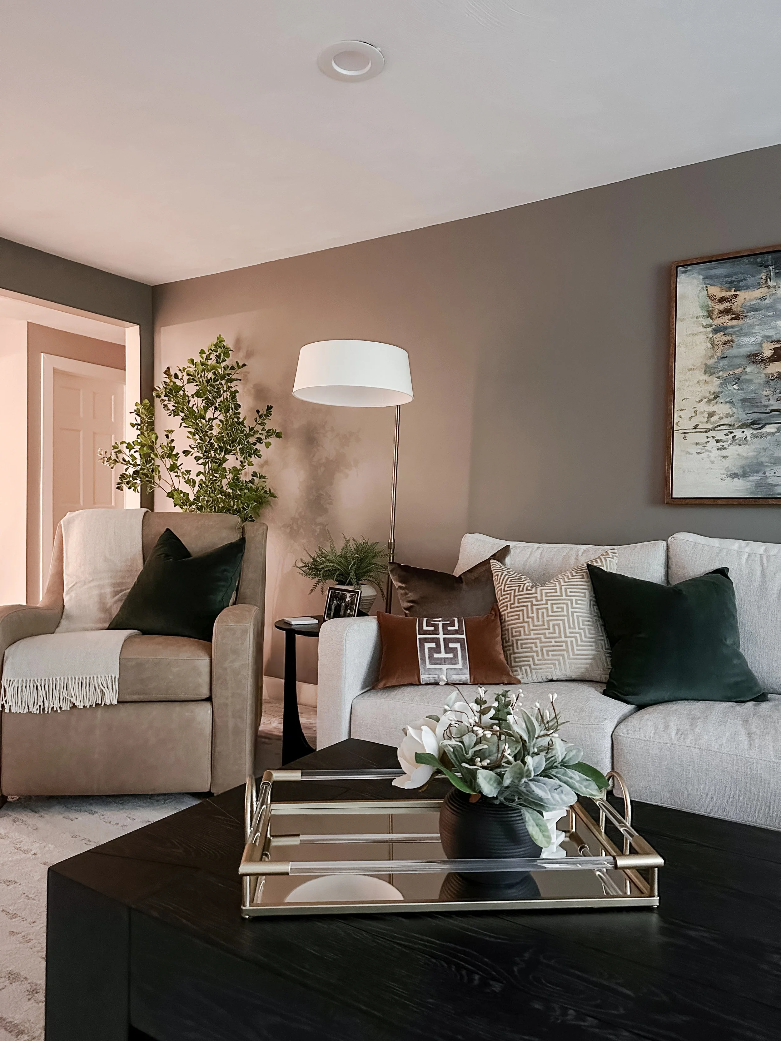 Living room with beige armchair, white sofa with patterned and solid colored pillows, black side table with a picture frame and plant, lamp, painting, and a coffee table with a vase of flowers.