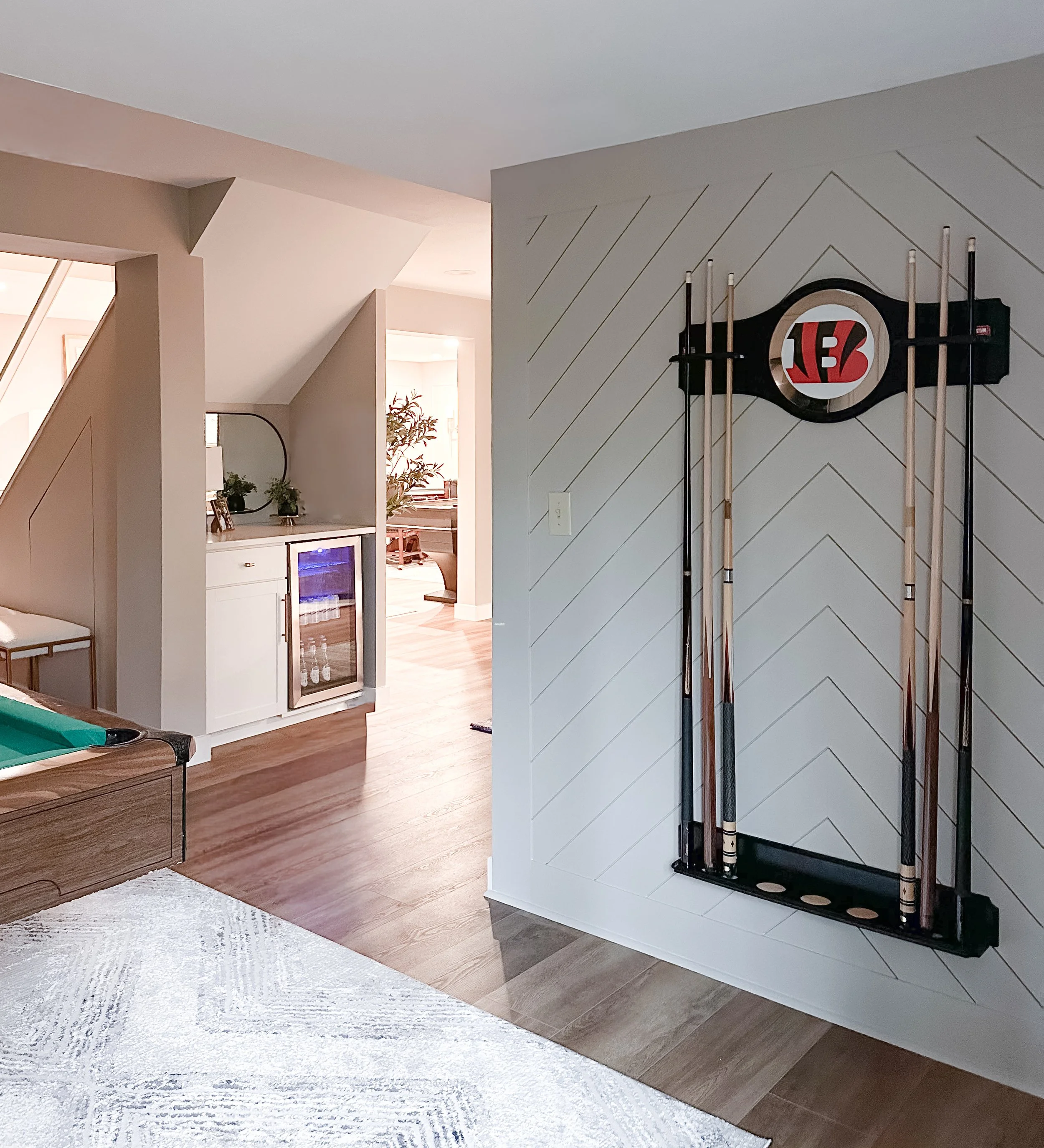 Indoor recreational area with a wall-mounted pool cue rack holding four pool cues, a Cincinnati Bengals logo on a round shelf, a small bar fridge, and a visible part of a pool table.