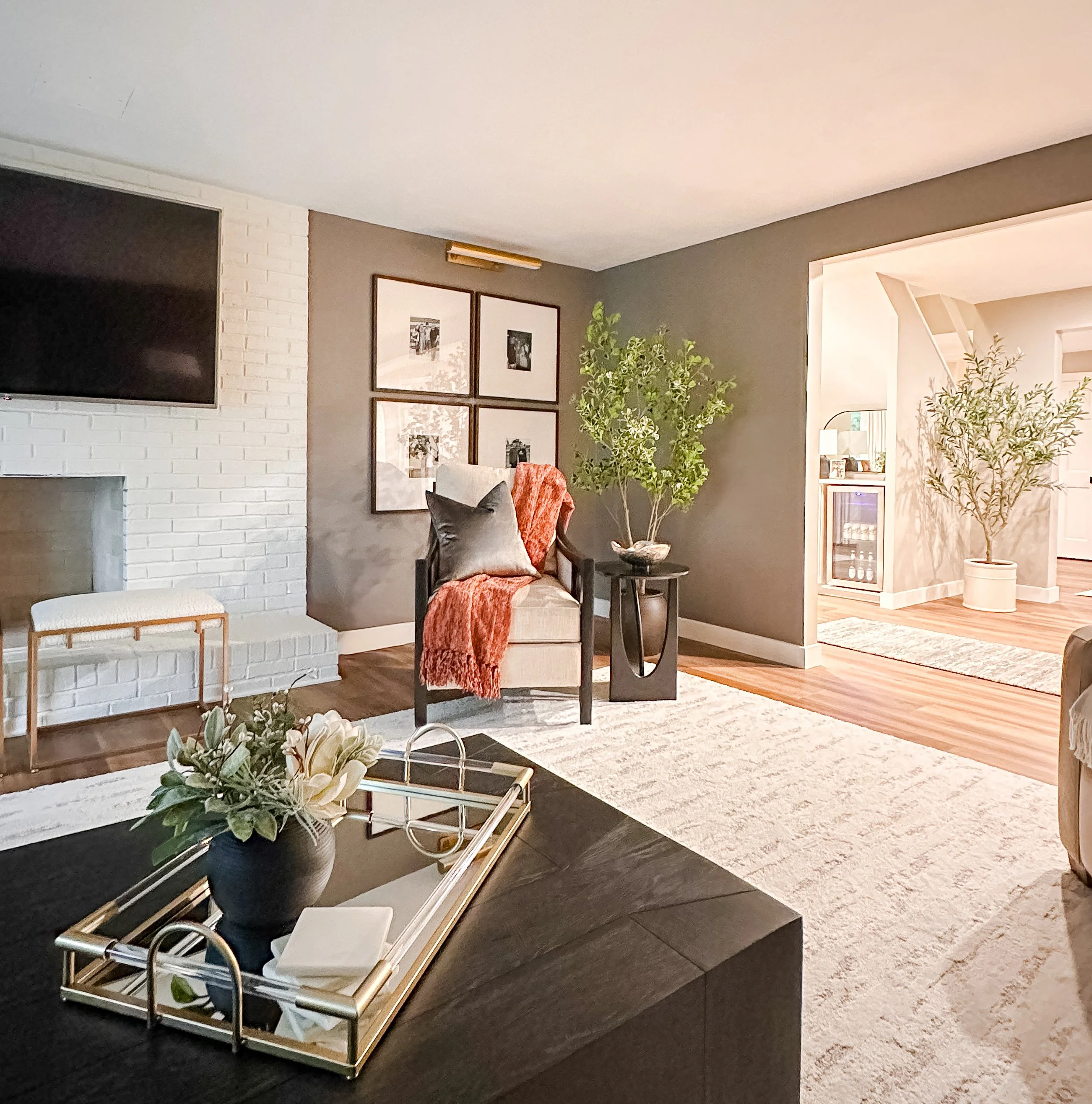 Living room with a gray accent wall, framed black-and-white photos, a beige armchair with a gray pillow, a red throw blanket, a black side table with a potted plant, a white brick fireplace, and a gold-framed tray with a flower arrangement on a black