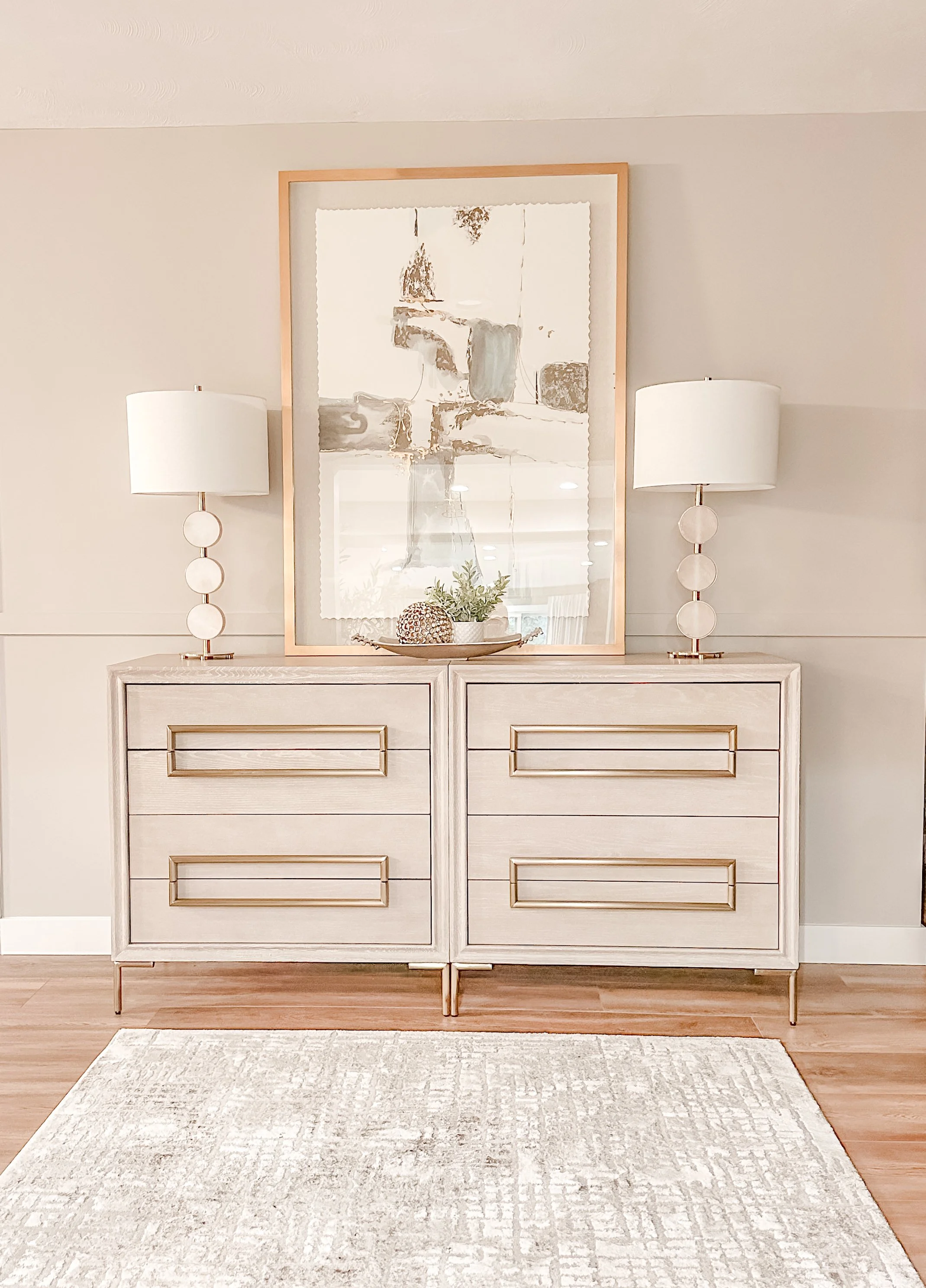 A modern console table with two lamps on either side, a large mirror above, and decorative items such as a plant and a decor ball, set against a neutral wall in a well-lit interior.