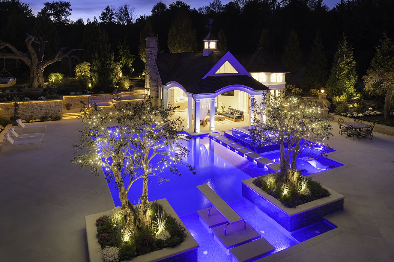 Nighttime view of a luxurious backyard with a lit swimming pool, illuminated trees with string lights, outdoor seating, and a well-lit house with a small porch.