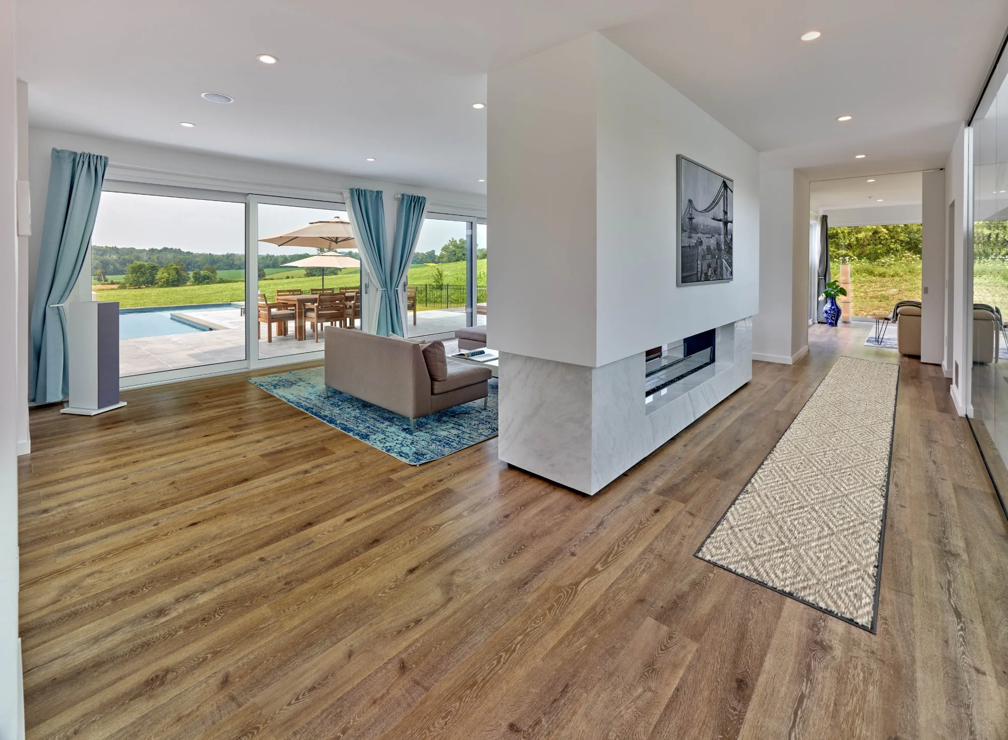 Modern living room with wooden flooring, a beige sofa, and a blue rug. Large sliding glass doors open to a backyard with a pool and outdoor dining area. A white fireplace or wall feature with a mounted black-and-white photo and a patterned runner rug on the floor.