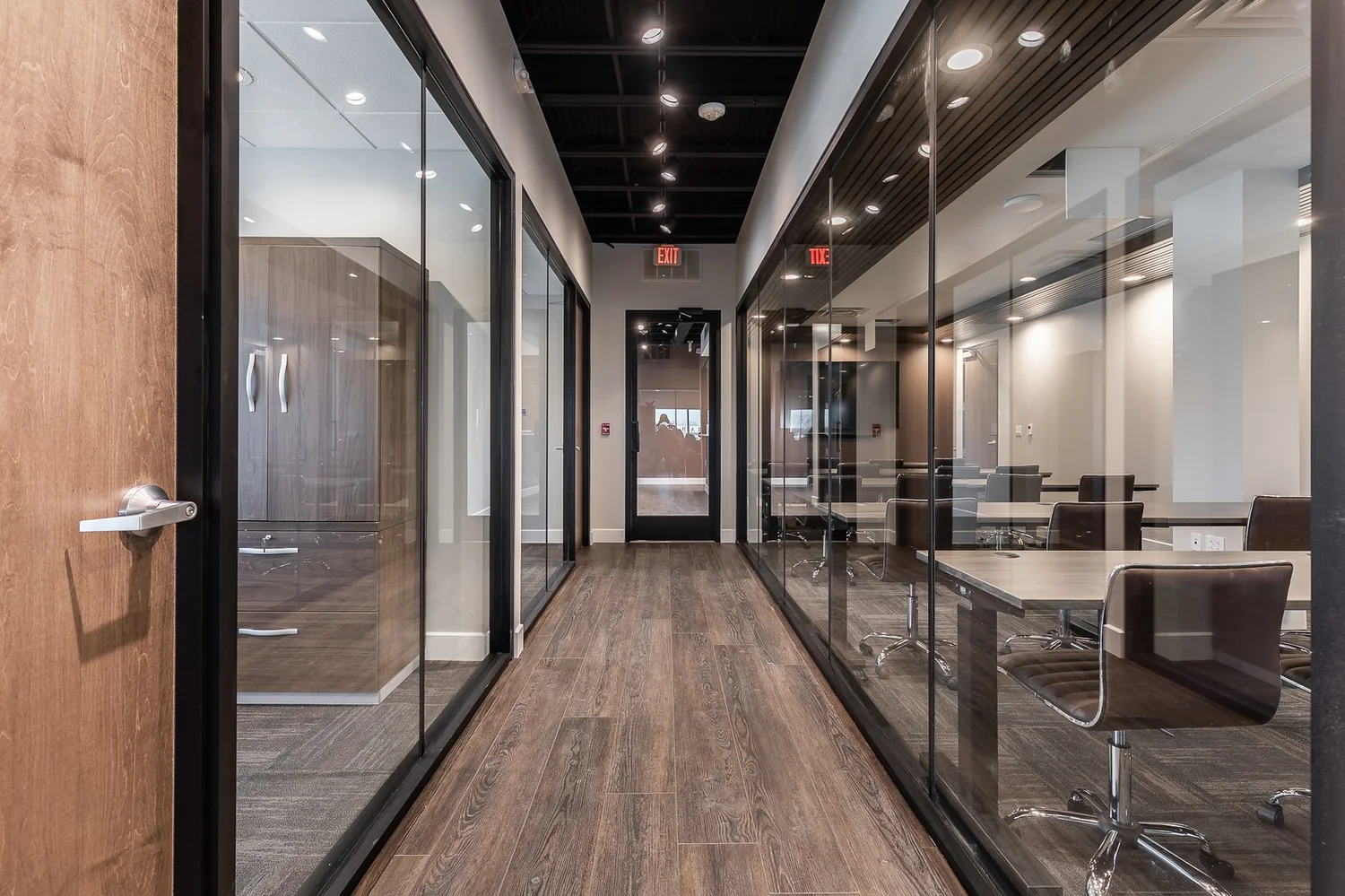 Office corridor lined with full-height glass walls and black frames, with wood-look floors and an open black ceiling