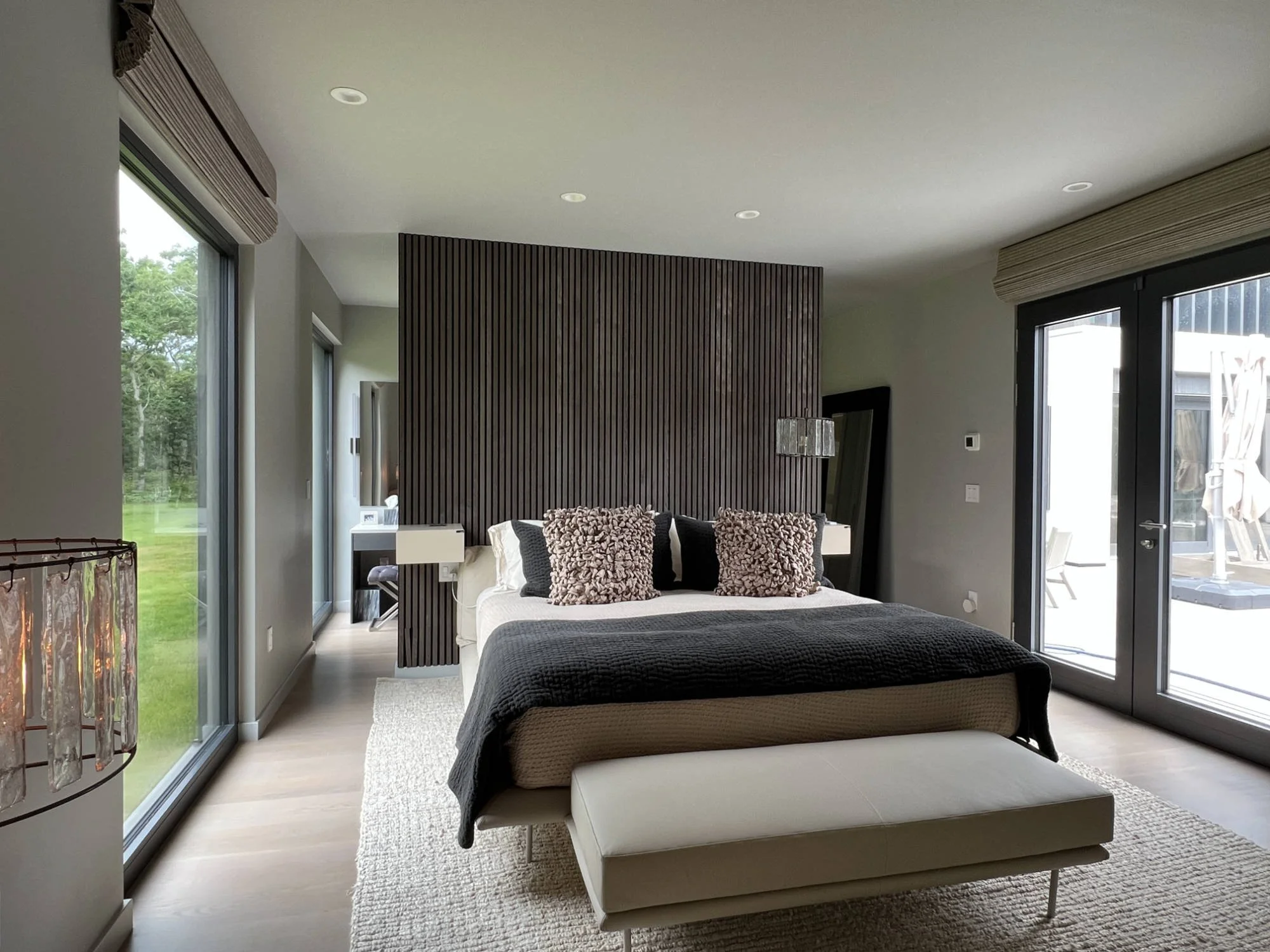 Modern bedroom with large glass sliding doors, a wooden headboard wall, a bed with decorative pillows and a bench at the foot of the bed, and a neutral color palette.