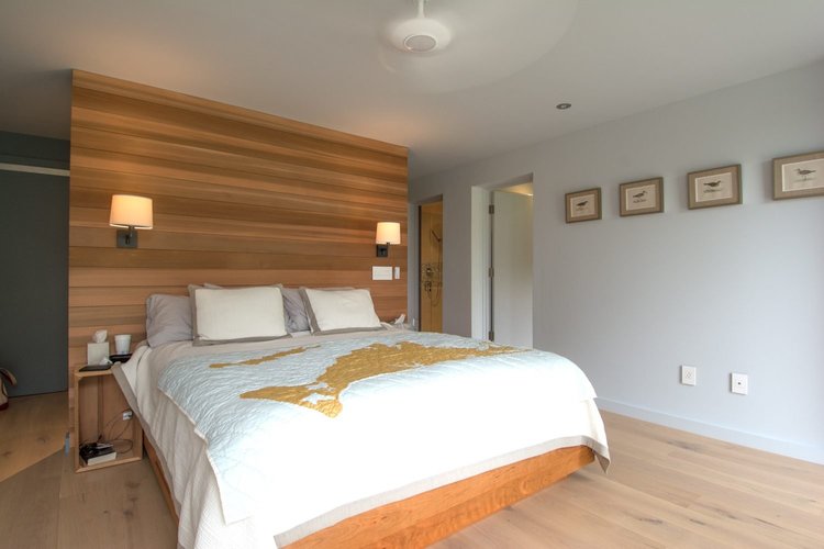 Bedroom on Martha’s Vineyard with a warm wood headboard wall, wall-mounted reading lights and framed prints