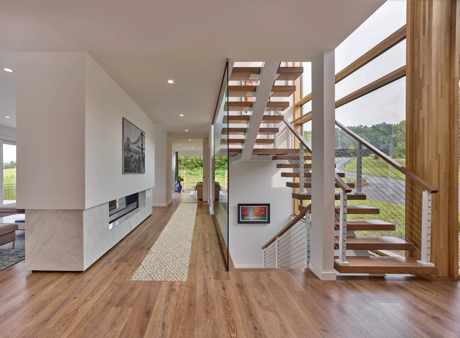 Long hallway with a wood floor leading to an open stair with wood treads, cable railings and tall wood fins along a window wall