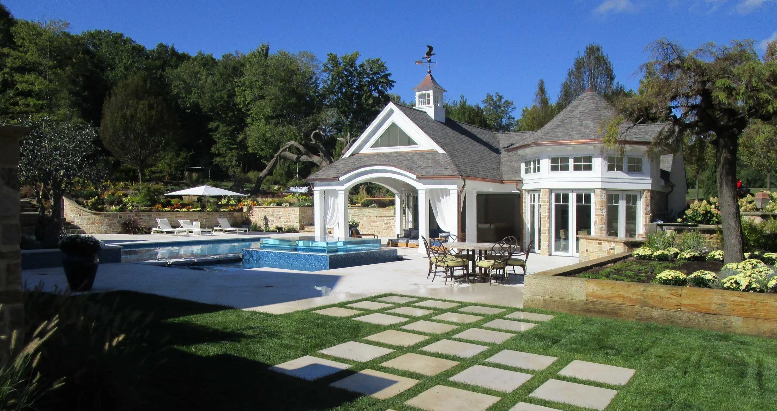 A backyard with a swimming pool, a round patio table with chairs, sun loungers with umbrellas, a stone house with large windows, a garden with flowers, trees, and a clear blue sky.