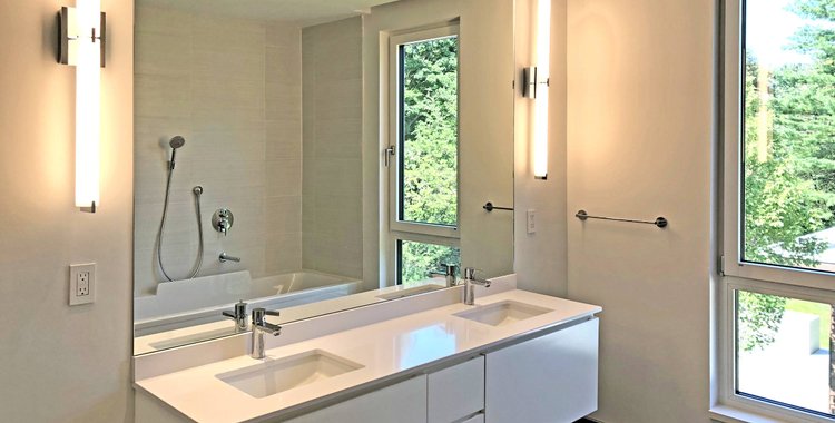 Bathroom with a long white double vanity, tall sconces beside the mirror and windows to the trees