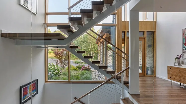Open stair with wood treads on a steel stringer, a cantilevered landing and cable railings in front of a two-story window to the garden
