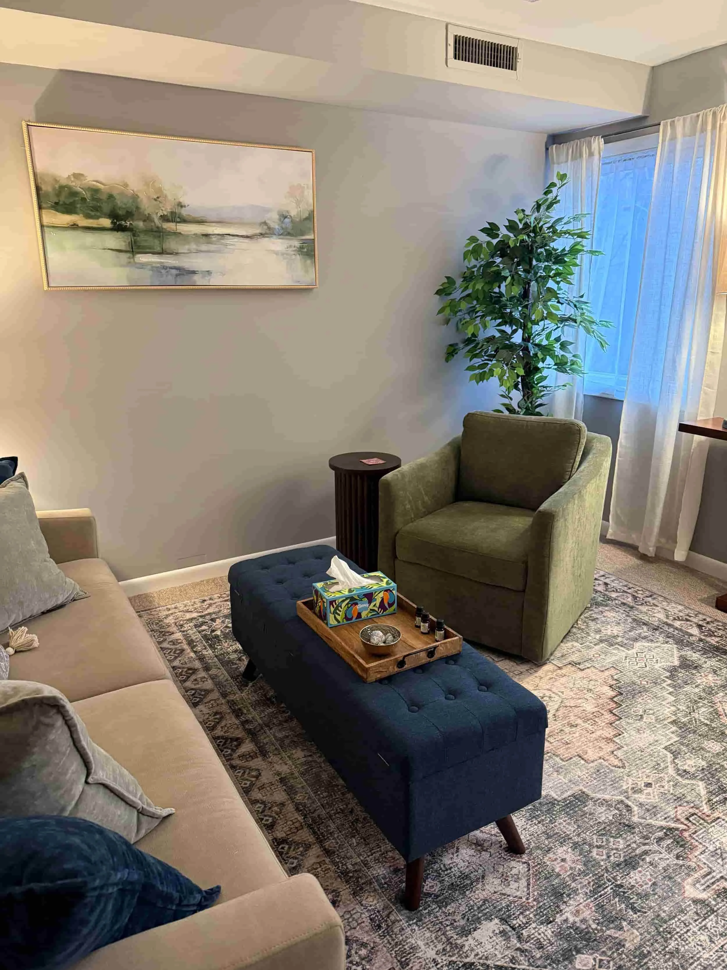 Living room with a beige sofa, green armchair, blue tufted ottoman, a wooden tray with tissues and small bottles, a patterned rug, a large wall painting of a landscape, a tall potted plant near the window with white curtains, and an air vent on the ceiling.