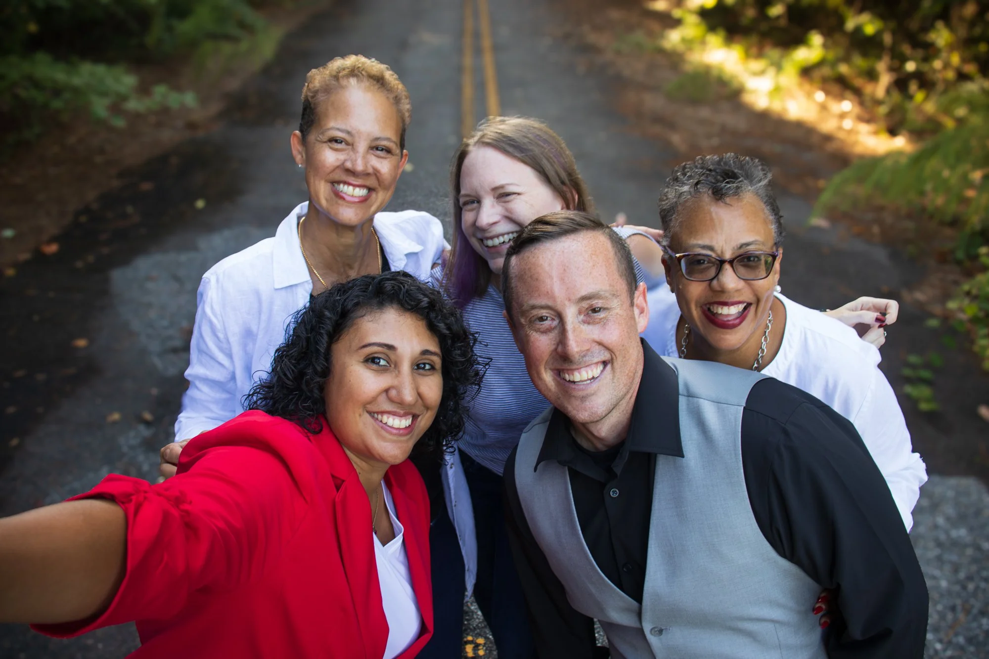 A diverse group of five smiling people taking a selfie on a road surrounded by trees.