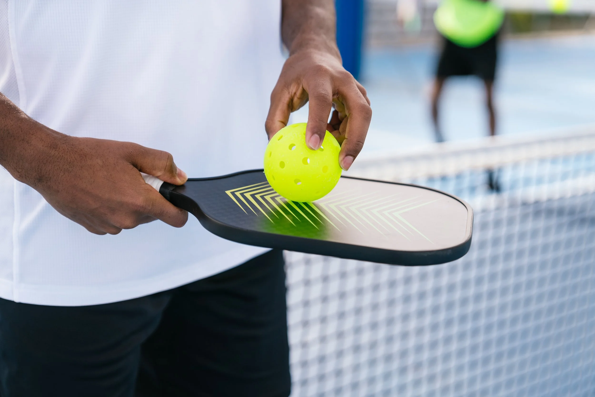 Person holding a paddle with a bright yellow pickleball on top during a pickleball game at the net, with a court and net visible in the background.