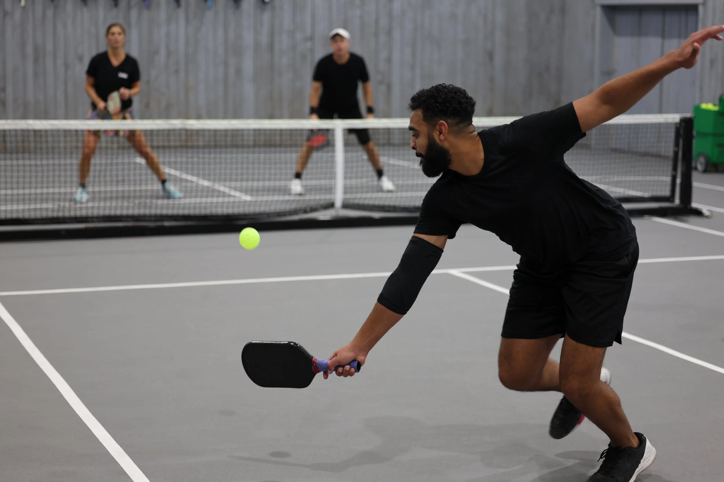 A man playing pickleball on an indoor court, preparing to hit a neon yellow pickleball with a paddle, with two women standing behind a net in the background.