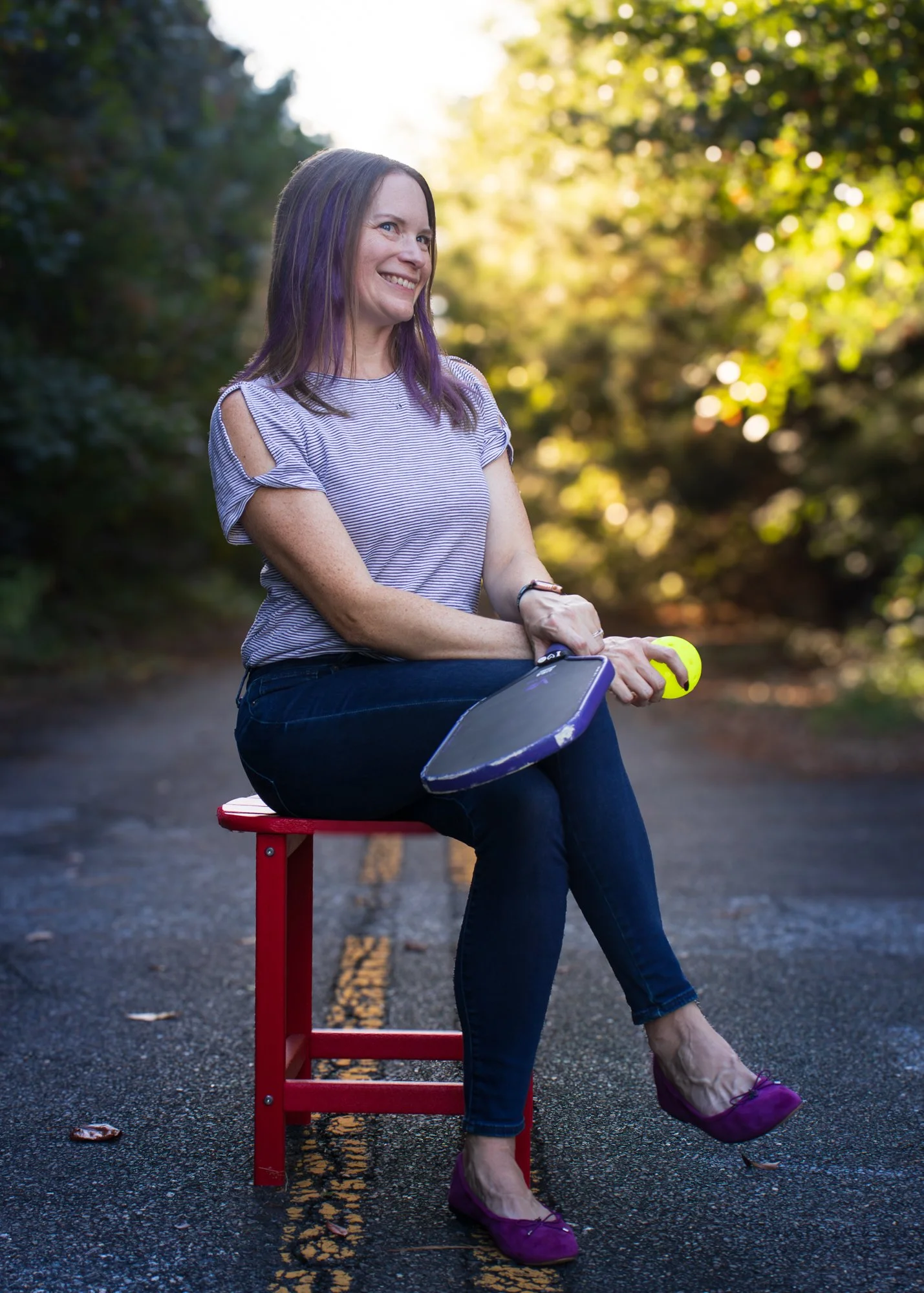 A woman with purple streaks in her hair, wearing purple shoes, is sitting on a red stool on a paved road with yellow lines. She is outdoors, smiling, with a background of green trees and sunlight.
