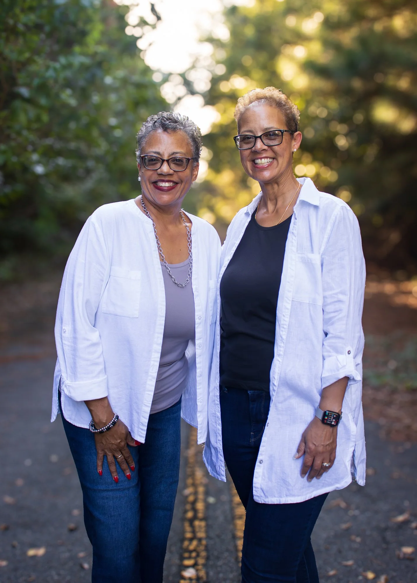 Two women standing outdoors on a tree-lined road, smiling at the camera. They are wearing glasses, white shirts over dark tops, and blue jeans. One has short gray hair, the other has short light brown hair.