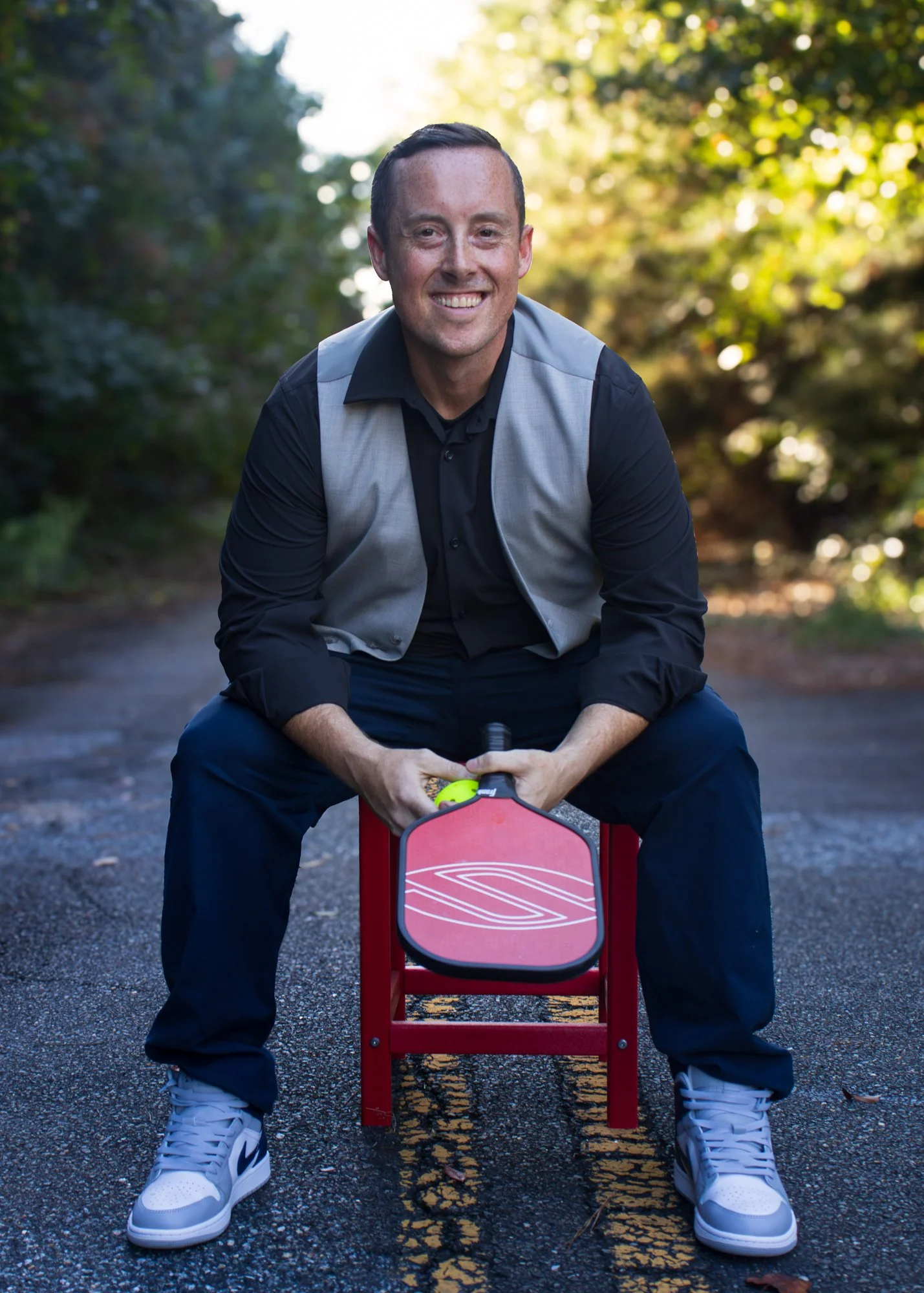 A smiling man sitting outdoors on a red chair, holding a pickle ball and paddle, with a bright background of trees and sunlight.