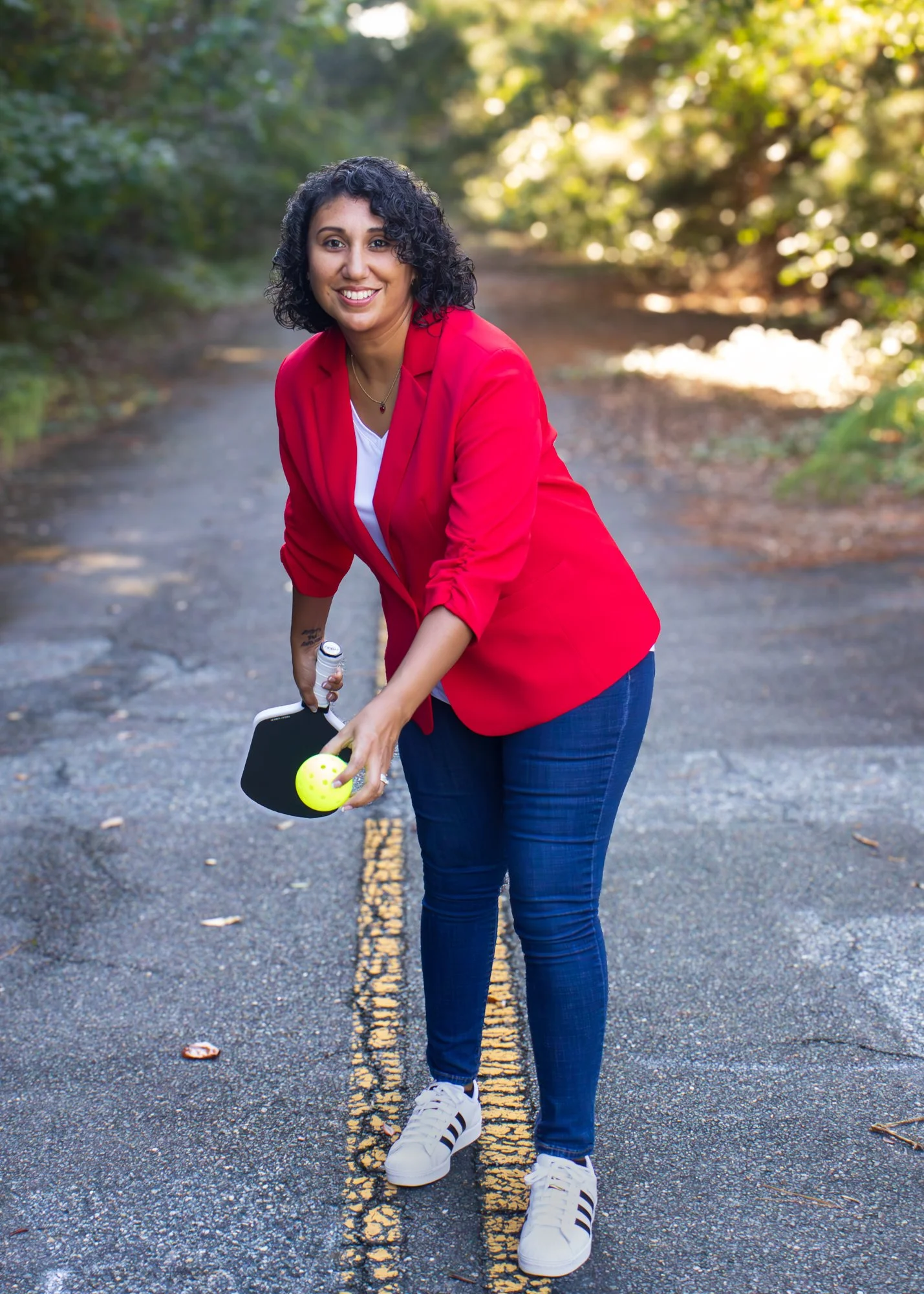 Woman in red blazer and jeans holding a yellow pickle ball and paddle standing on a rural road surrounded by trees
