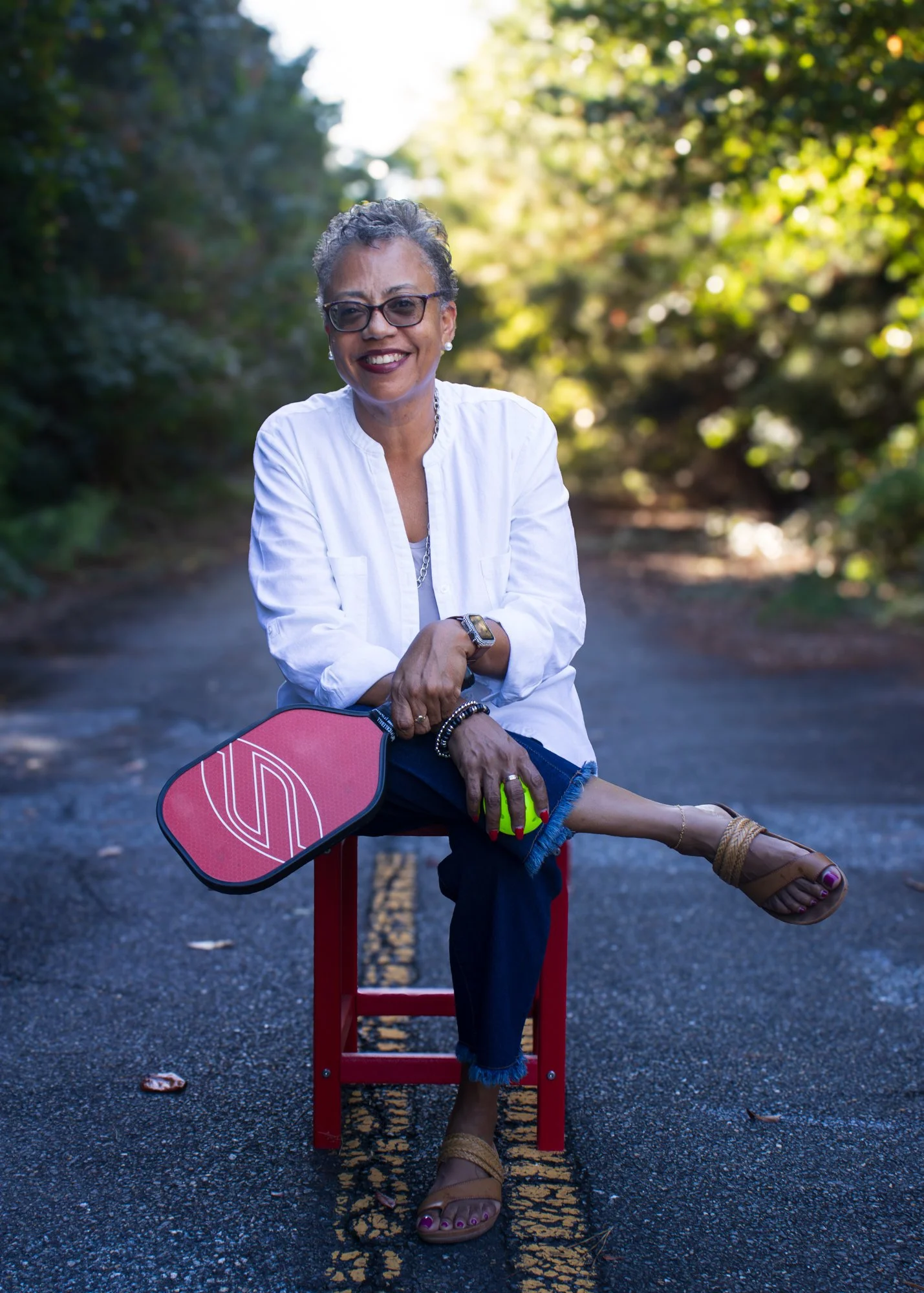 An older woman with short gray hair, glasses, and jewelry, sitting outdoors on a red chair on a paved road, smiling and wearing a white shirt, dark pants, and tan sandals, with lush green foliage in the background.