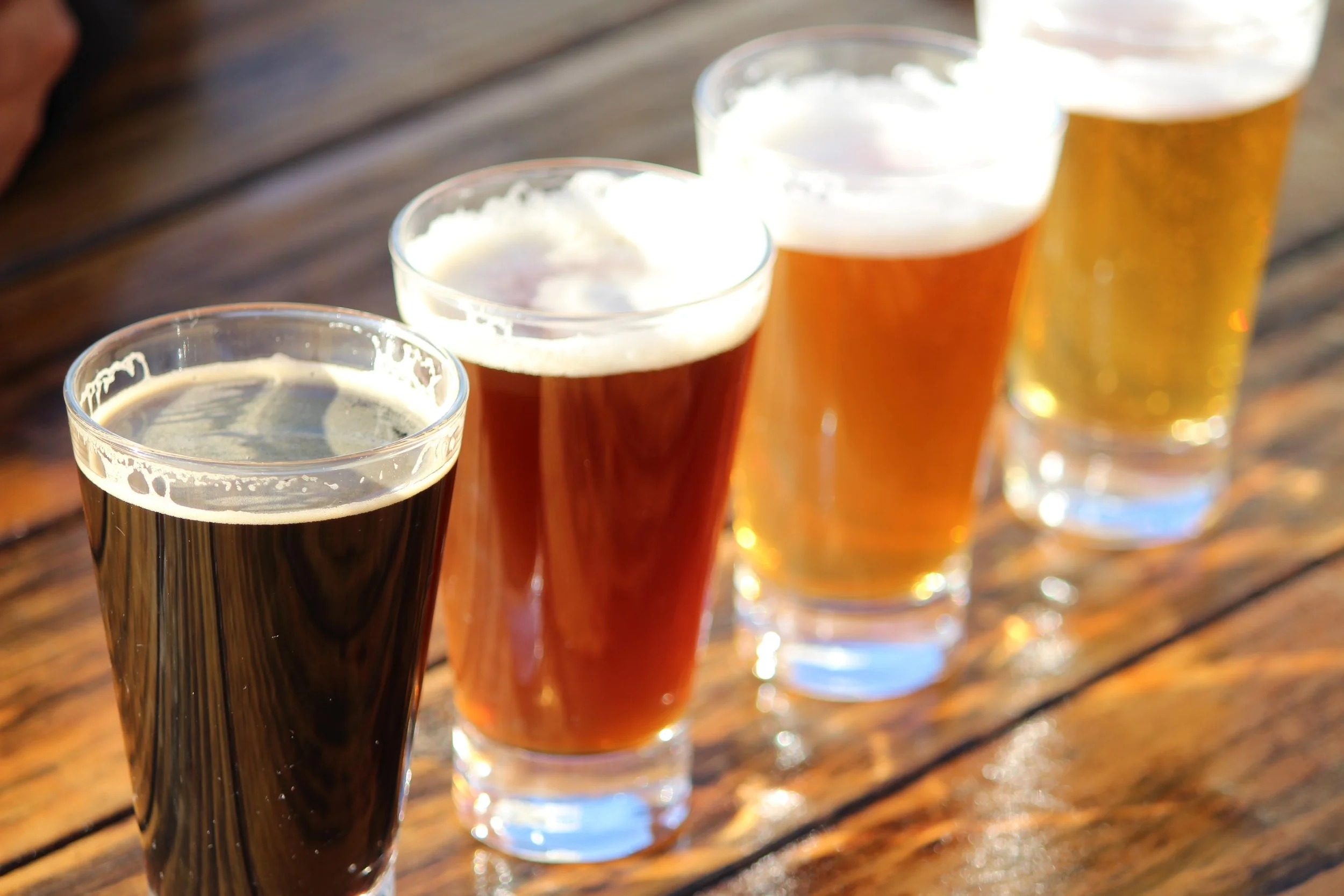Four glasses of beer with foam on top, lined up on a wooden table with sunlight reflecting.