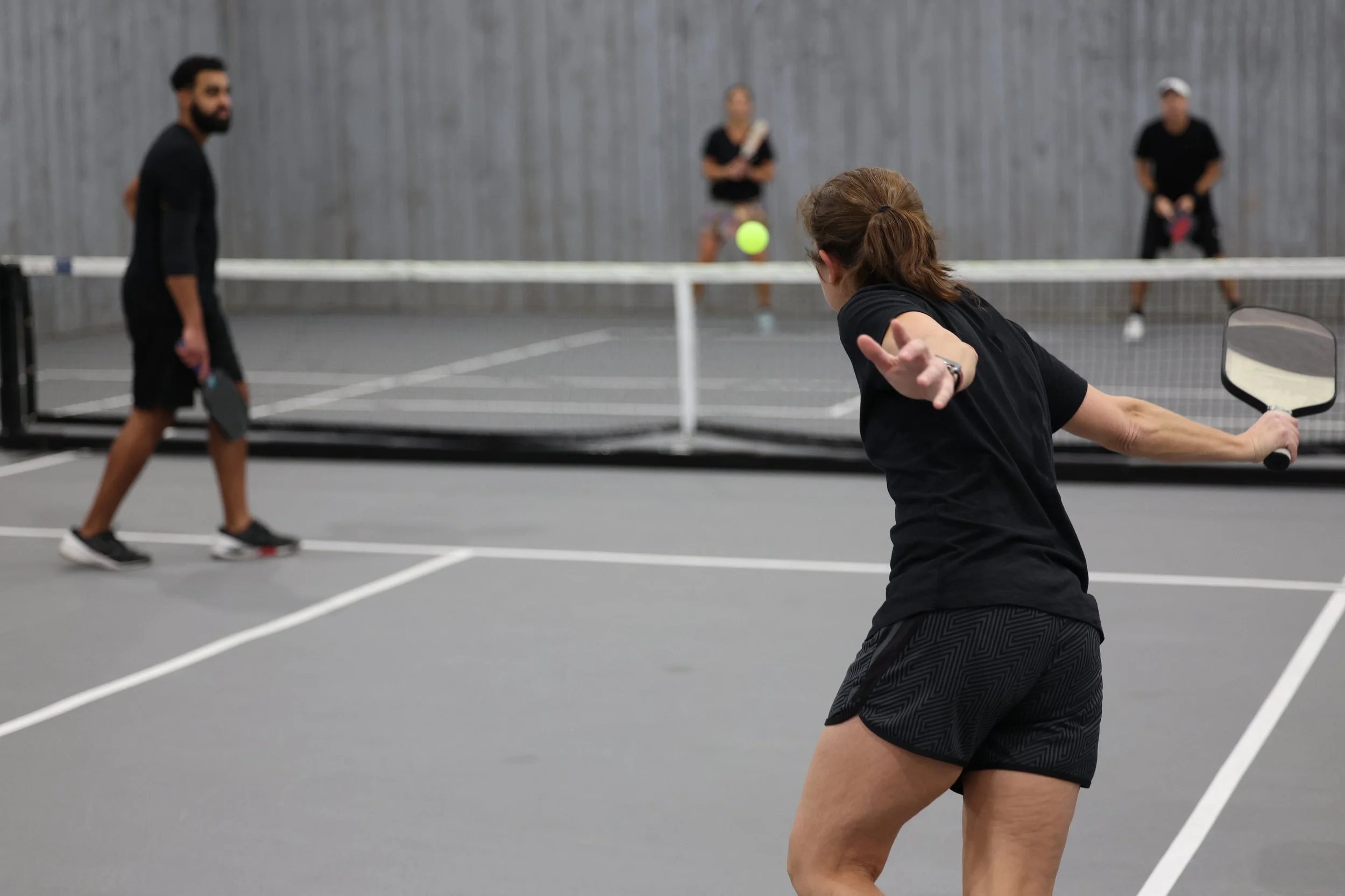 A group of people playing pickleball indoors on a grey court, with a woman in black shorts and shirt preparing to hit a green ball, a man holding a paddle on the left, and two other players in the background near the net.