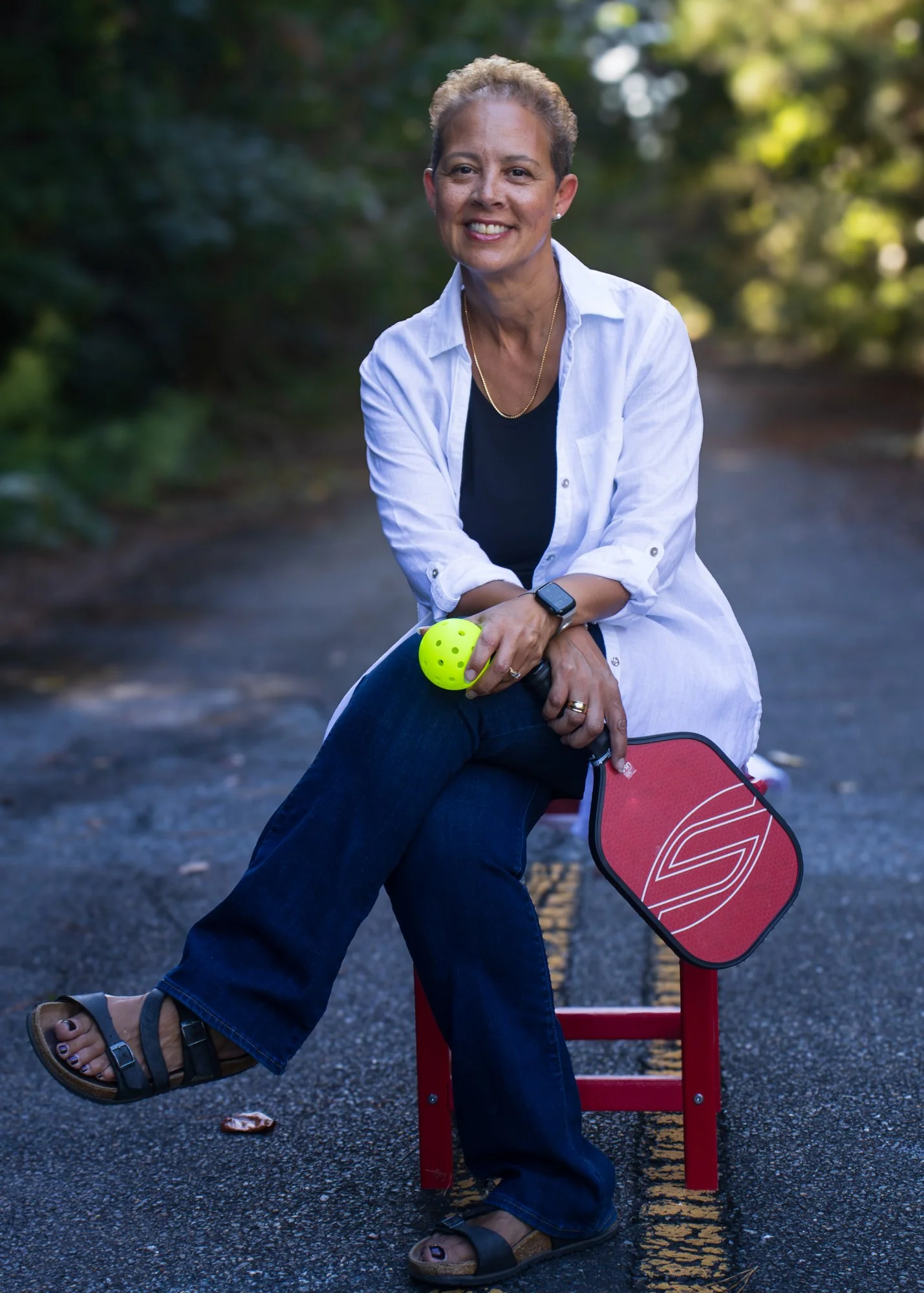 A smiling woman sits on a red bench outdoors, holding a green tennis ball, with a paddle next to her. She is wearing a white shirt, dark jeans, sandals, and a smartwatch, with short curly hair, in a green, leafy park setting.