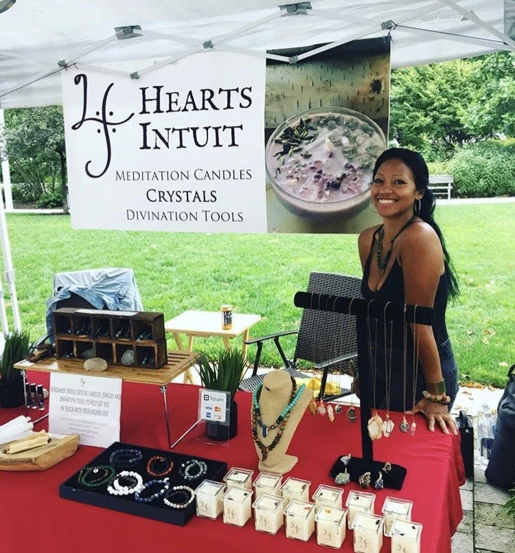 A woman standing behind a jewelry and crystal stand at an outdoor market, with a banner that reads "Hearts & Intuit, Meditation Candles, Crystals, Divination Tools." The table displays necklaces, bracelets, and small crystal boxes, with greenery and trees in the background.