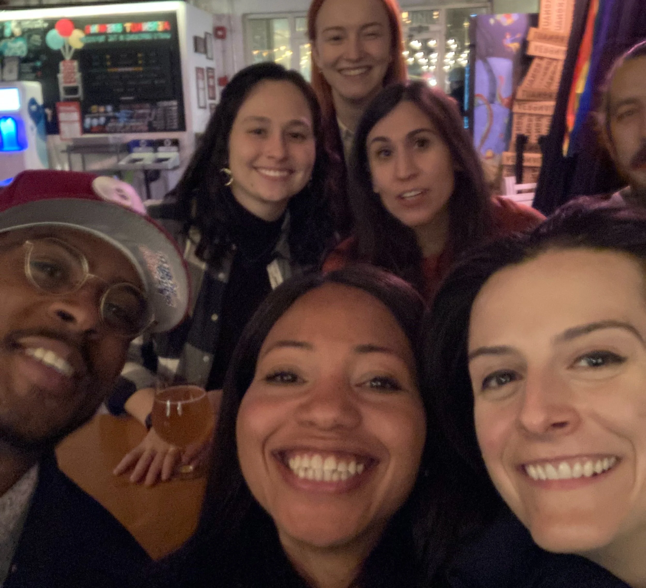 A group of diverse women smiling and celebrating together with drinks in a lively indoor setting, possibly a restaurant or bar, with colorful decorations and lights in the background.
