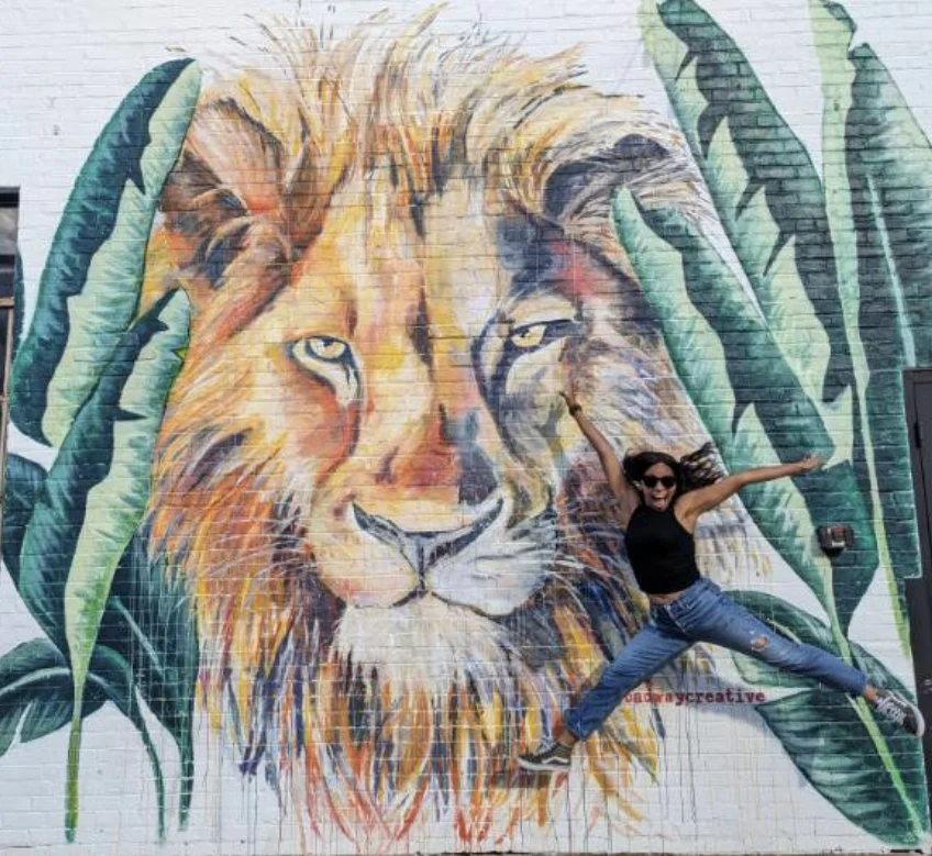 A woman jumping in front of a colorful mural of a lion's face on a brick wall, with large green leaves painted around it.