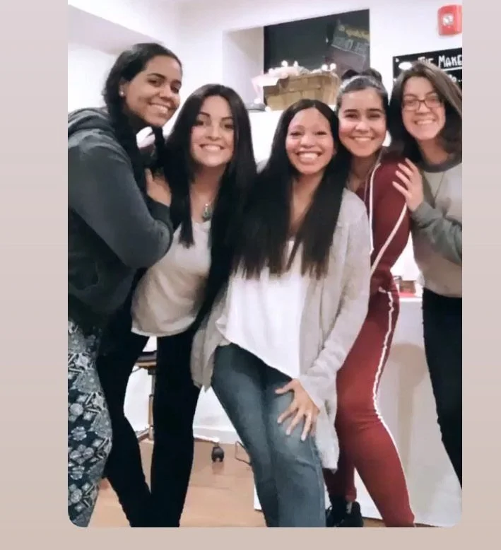 Five women standing close together, smiling, inside a room with a chalkboard and some shelves in the background.