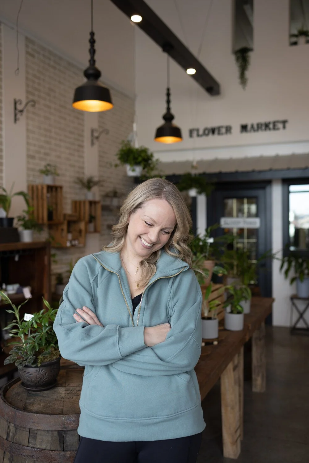 A woman smiling with crossed arms at a plant shop named Flower Market, surrounded by various potted plants, wooden shelves, and hanging lights.