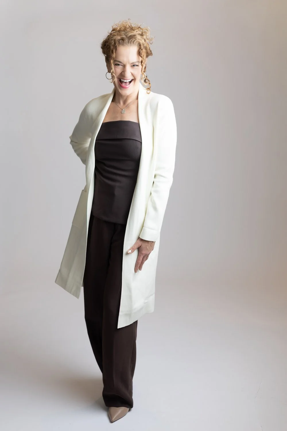 Photographer and coach Maggie Nowak, owner of Story Within wearing a white coat over a black outfit, smiling and laughing against a plain gray background.