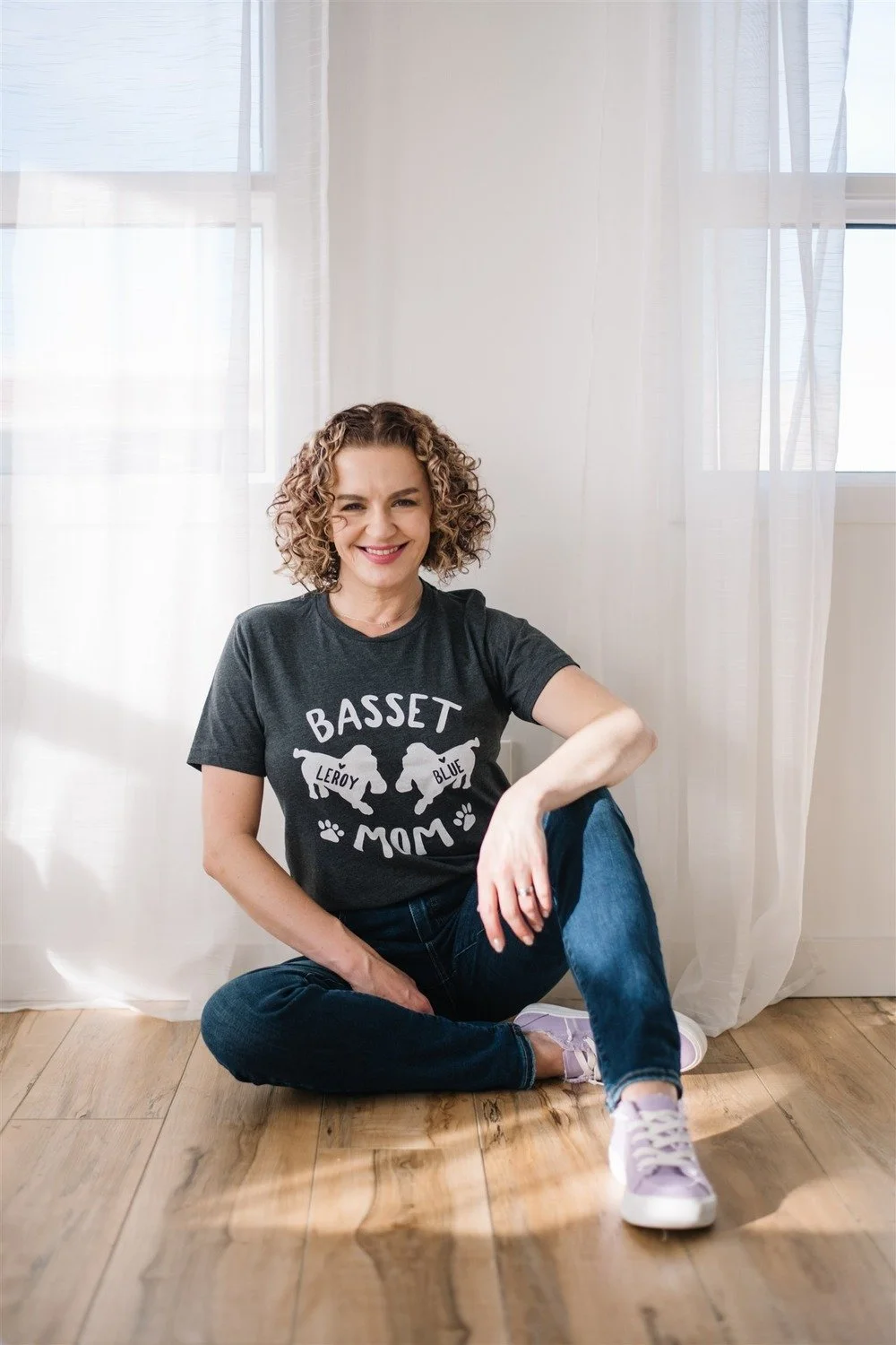 Photographer and coach Maggie Nowak, owner of Story Within, sitting on the wooden floor beside a window with sheer curtains.