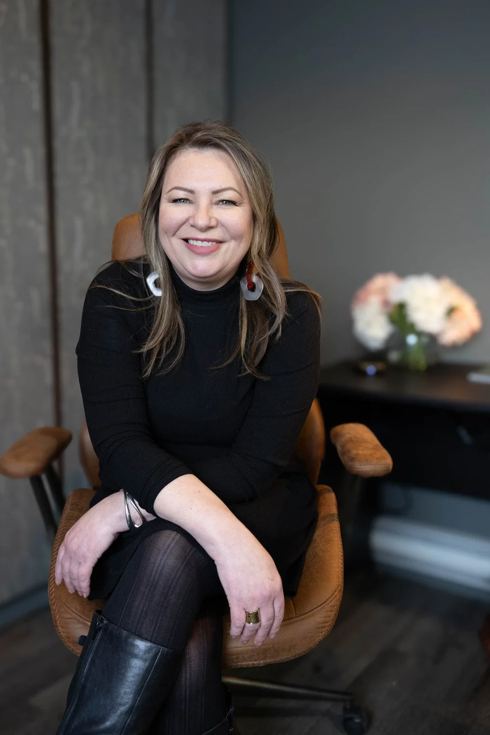 A woman with long hair, wearing a black top and large earrings, sitting in a brown chair, smiling at the camera.
