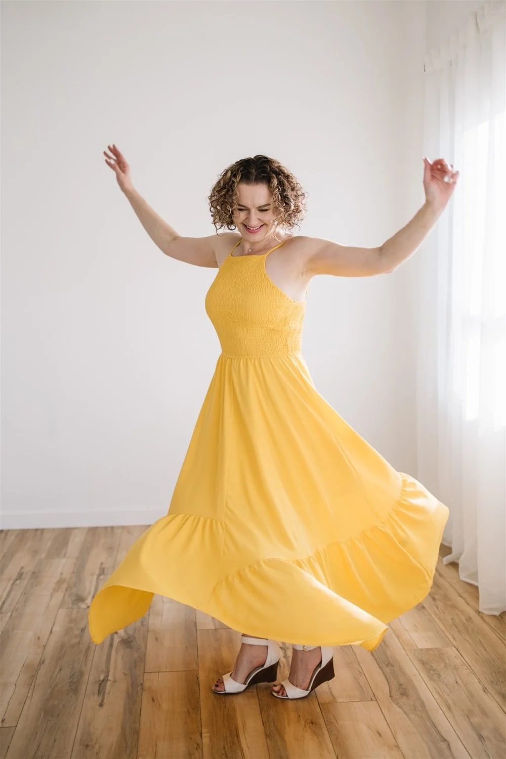 Owner of Story Within, Maggie Nowak, dancing in a yellow dress and white high heels in a room with wooden floors and white walls.