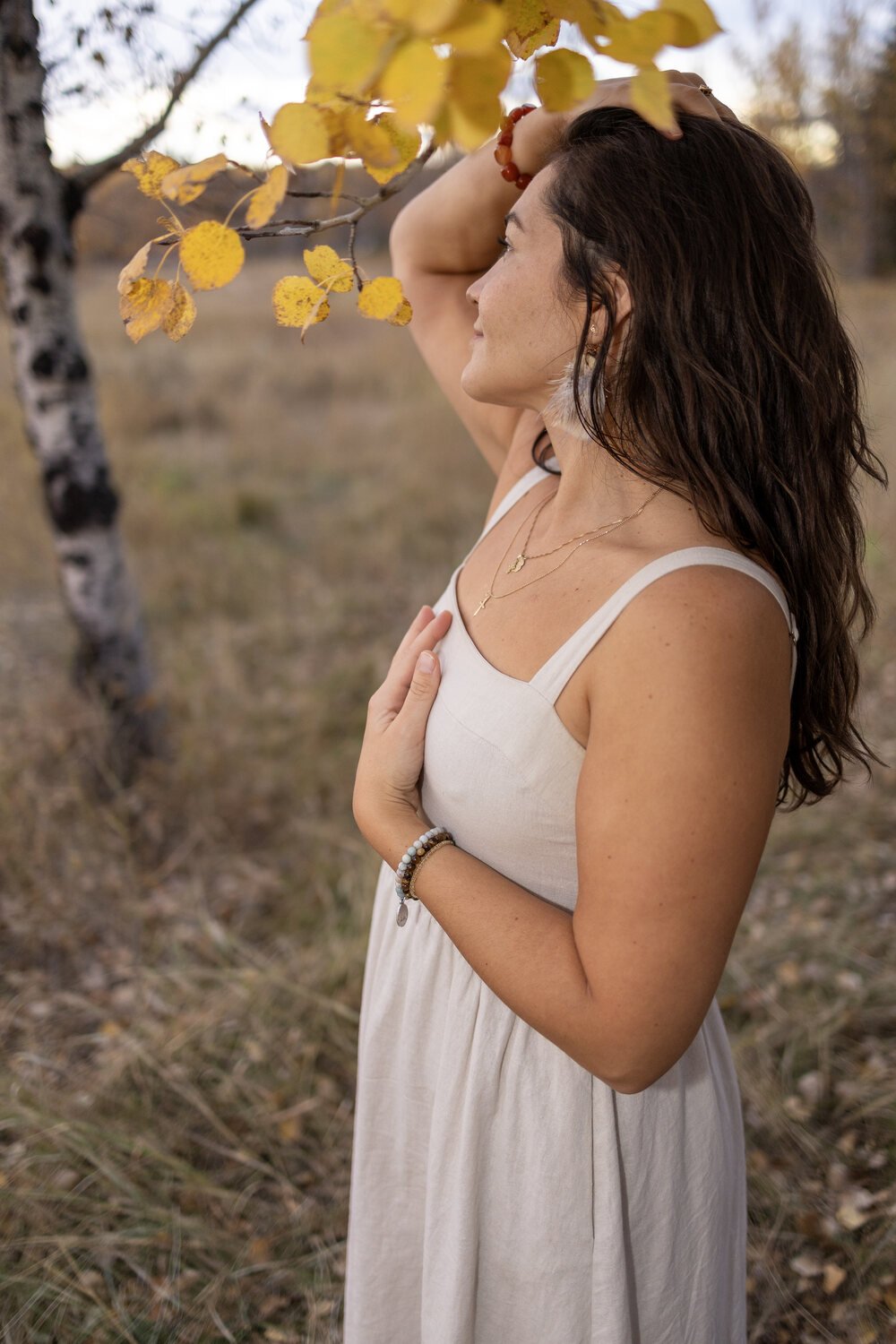 A woman with dark, wavy hair, wearing a white dress and layered necklaces, stands in a field with trees and yellow leaves, touching her chest and holding her head with one hand, captured during autumn.